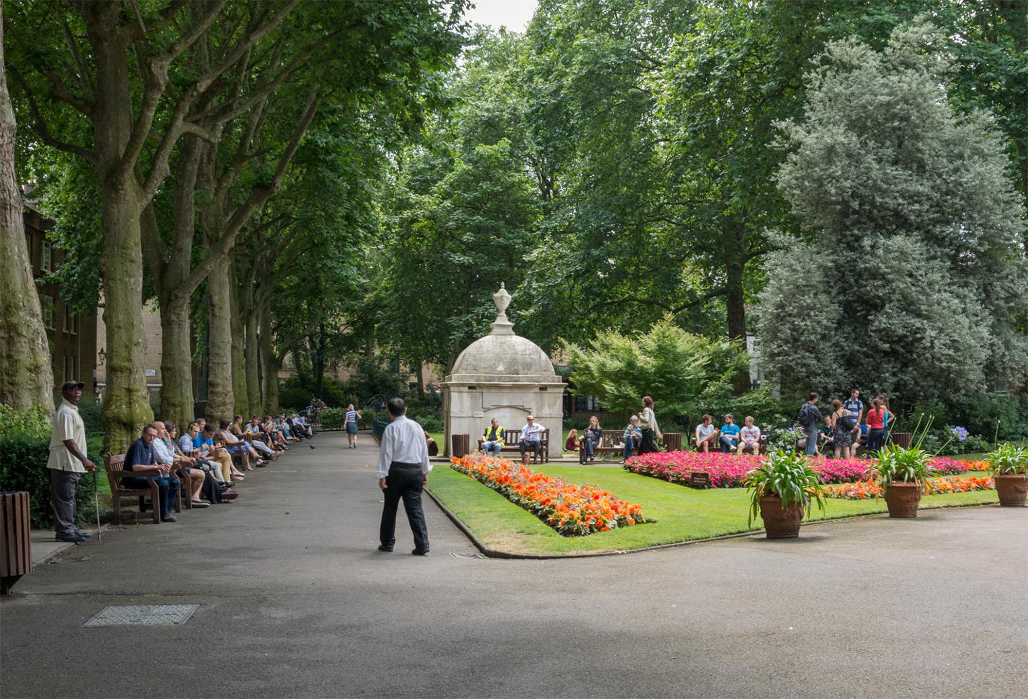 People seated on benches around flower beds in a park with a mausoleum in the background