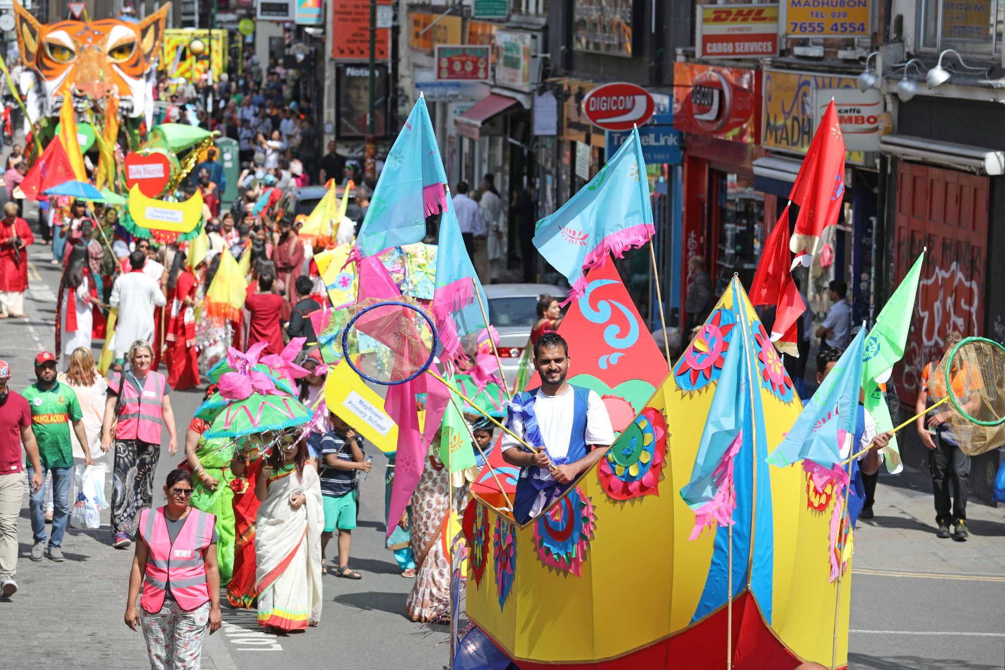A carnival parade makes its way up a road. People wear colourful clothing, and carry flags. At the head of the procession a man walks inside a boat made of fabric stretched around a frame.