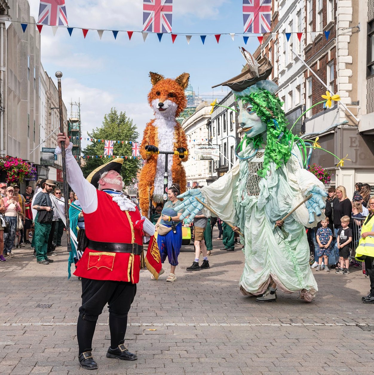 A carnival street scene with a town crier character in the foreground and large puppets and crowds in the background.