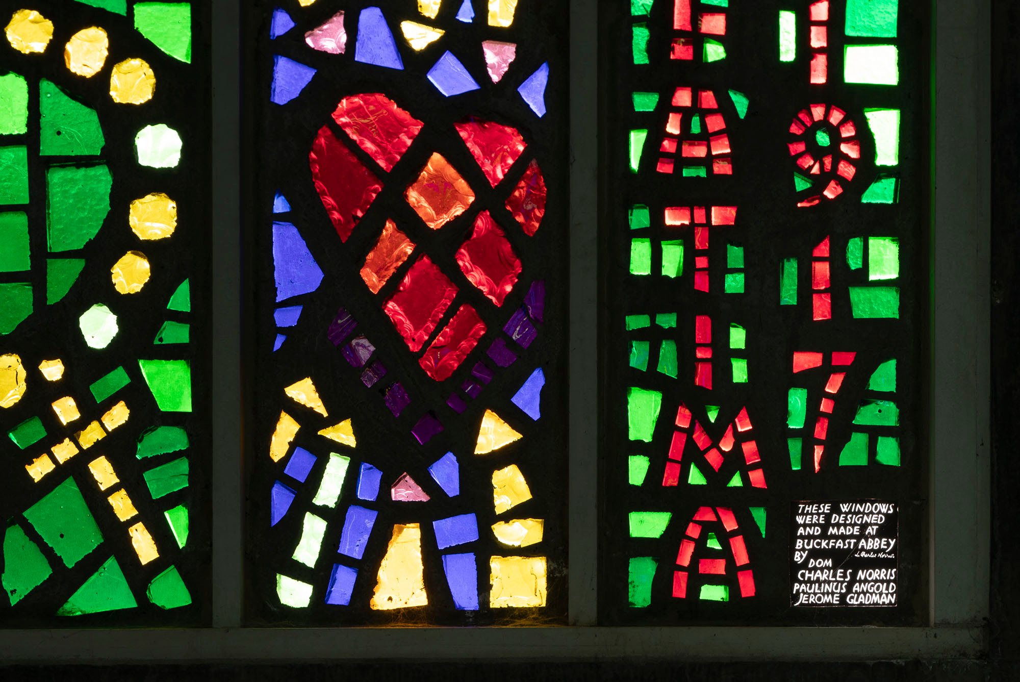 The photo shows three panels of chunky glass design, with vibrant red, green, yellow and blue in a black background, with a heart in the centre panel.