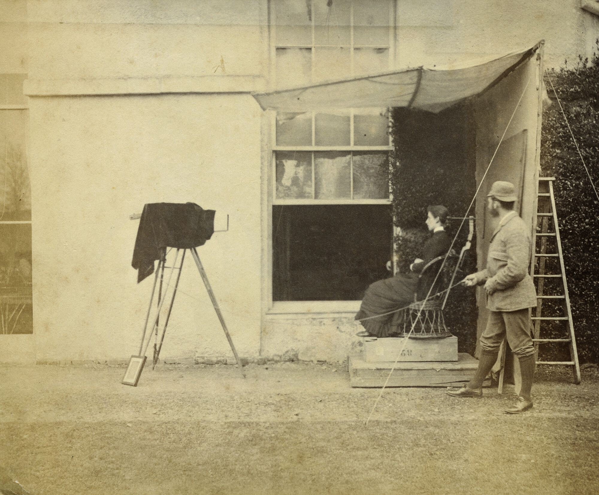 A whiskered photographer, in jacket, knickerbockers and deerstalker (far right), holds a cable attached to a camera mounted on a tripod. His subject, a young woman sits in a chair raised by two plinths. This studio is set up outside a house and adjacent to an open sash window and wall covered with foliage.
