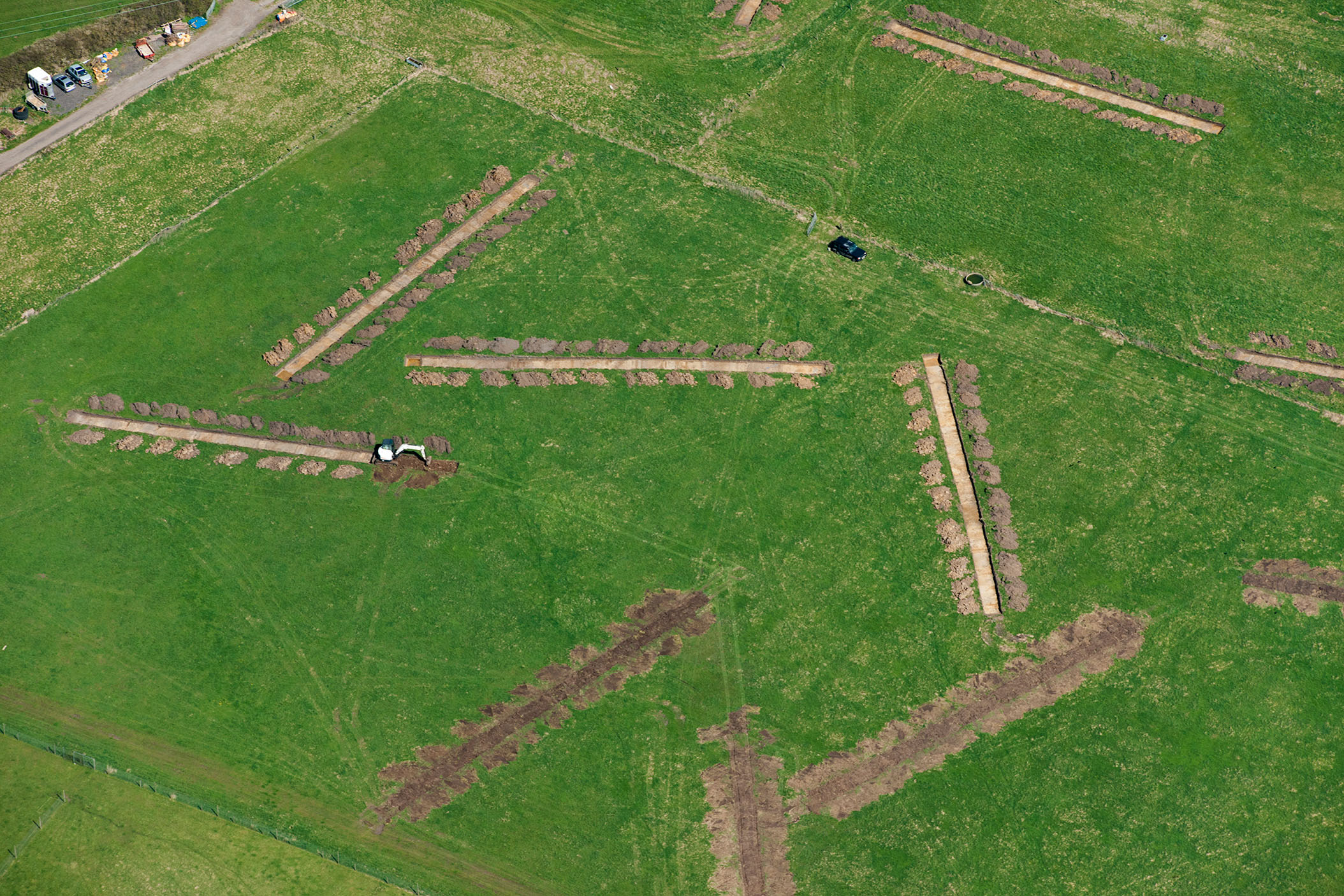 An aerial photograph of eight trial archaeological trenches in a green field.