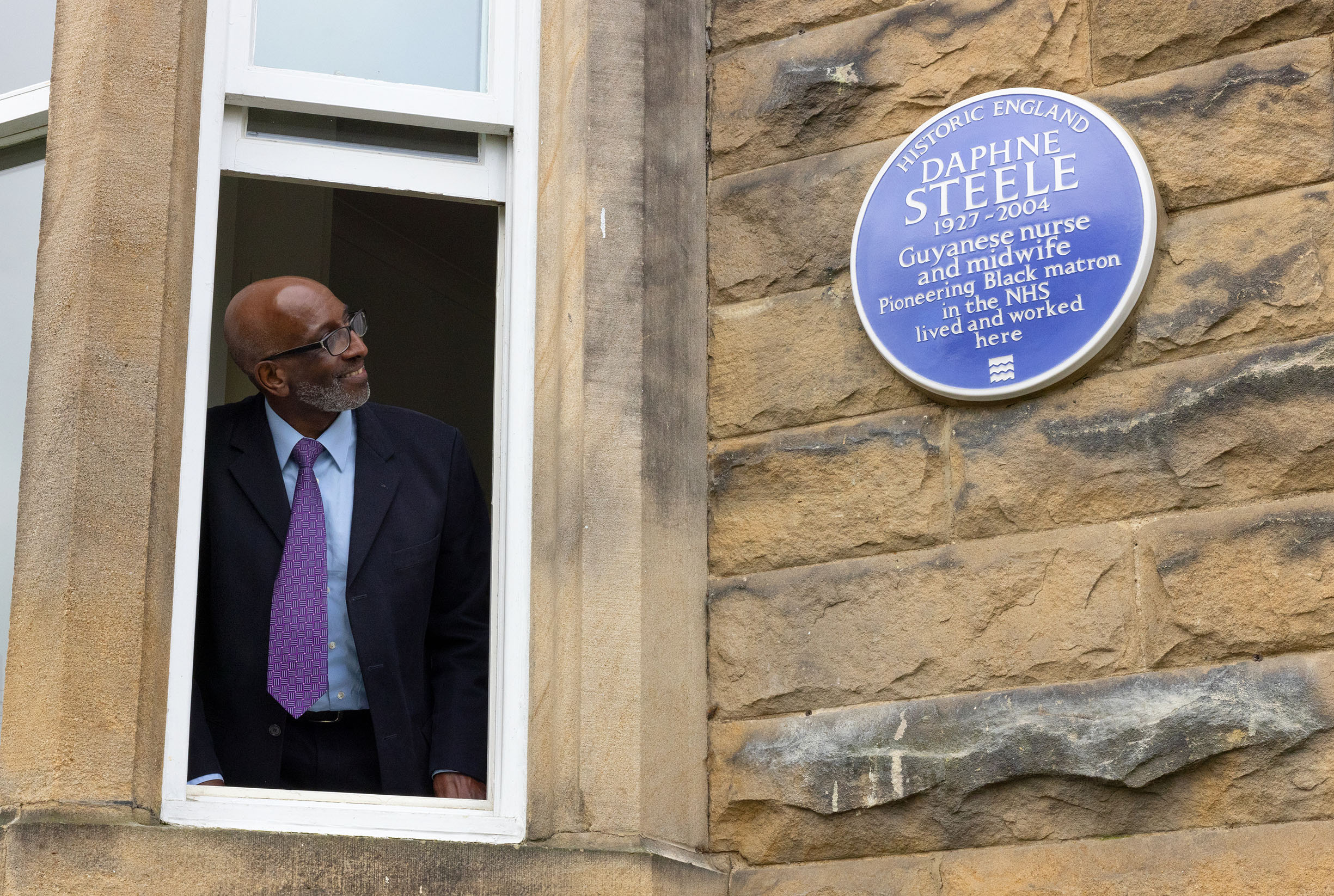 A man in a suit and tie stood at an open window, turned to look at a heritage blue plaque on a wall next to him