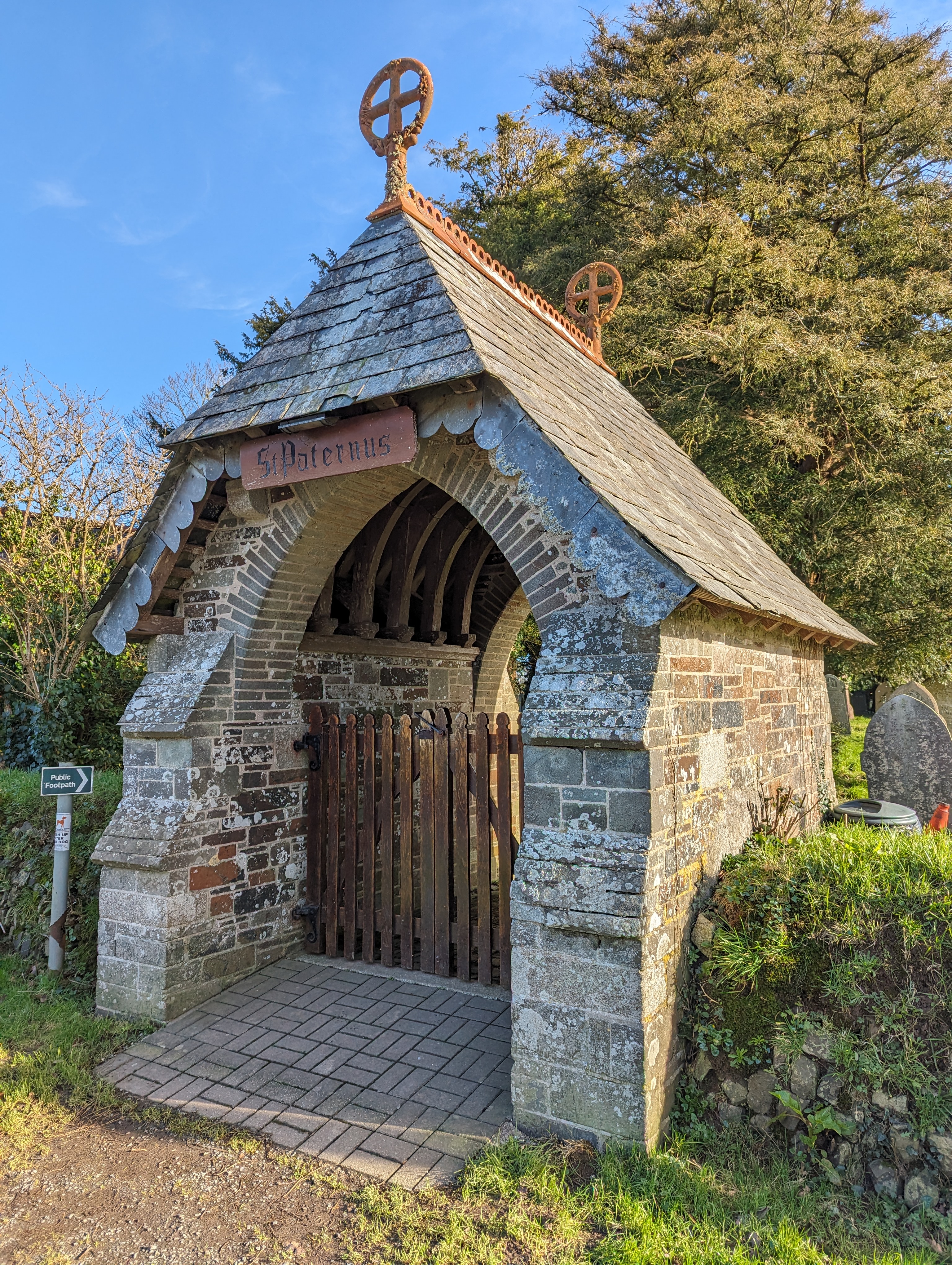 A portrait photograph of a stone entrance to a churchyard with a wooden gate and sloped tiled roof. A wooden sign above the entrance reads "St Paternus"