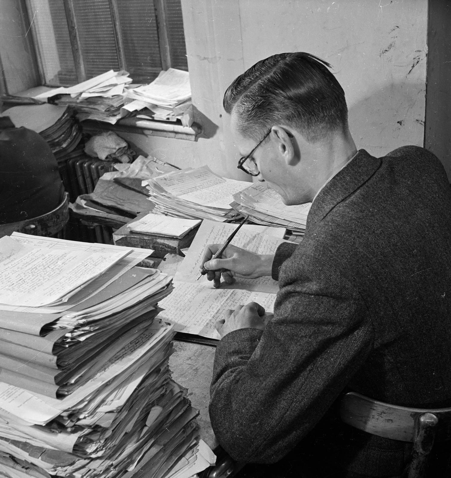 A man wearing a suit writing at desk surround by piles of paper