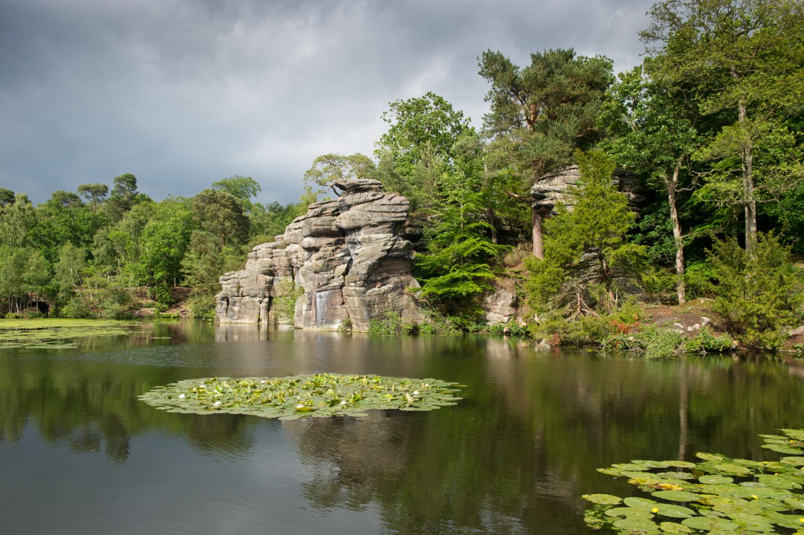 Lake with water lillies in the foreground bordered by dramatic rock features and trees.