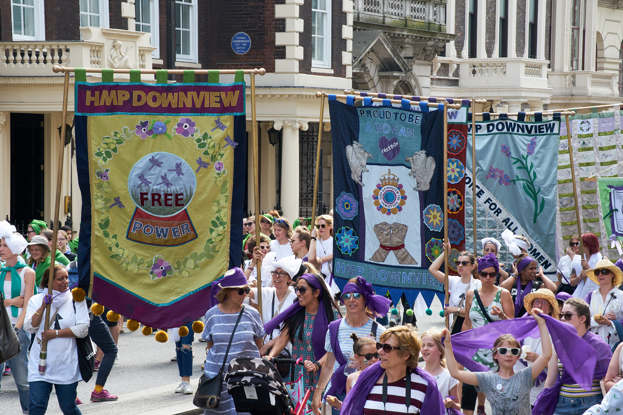 Three of the banners are marched down Whitehall by women and girls wearing green white and purple.  The banners have a mixture of empowering statements such as ‘HMP Downview Free Power’, ‘Proud to be a woman’ ‘Votes for all women’.