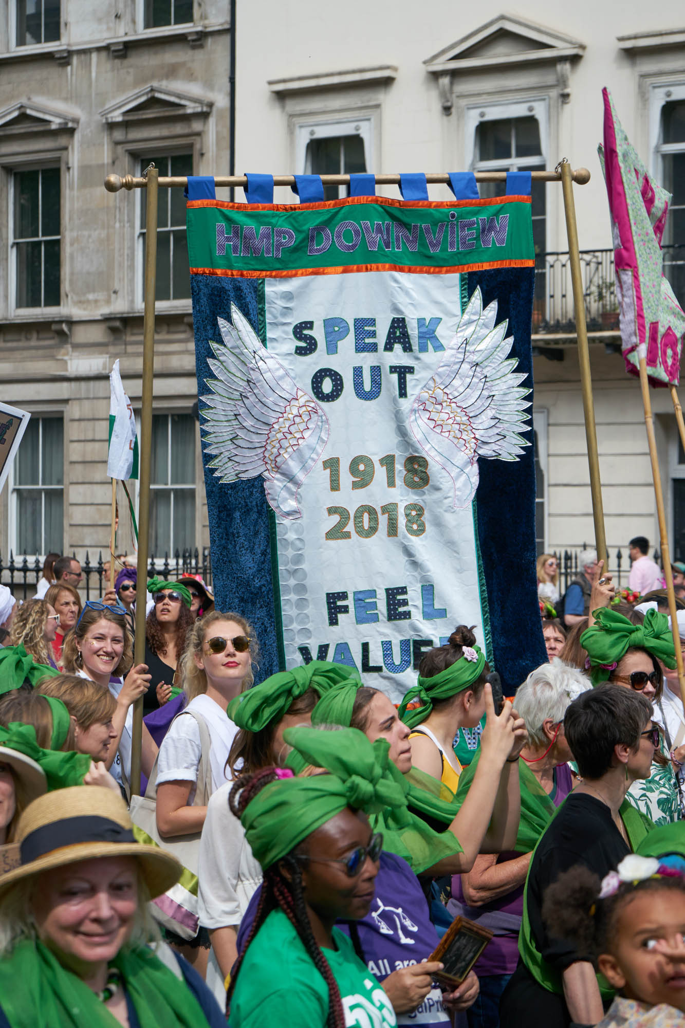 A green blue and white banner with the words ‘Speak out 1918-2018 Feel valued’ in green on the white middle section.  A pair of angel’s wings have been attached to it  A sea of women wearing green hold the banner as they walk down Whitehall.