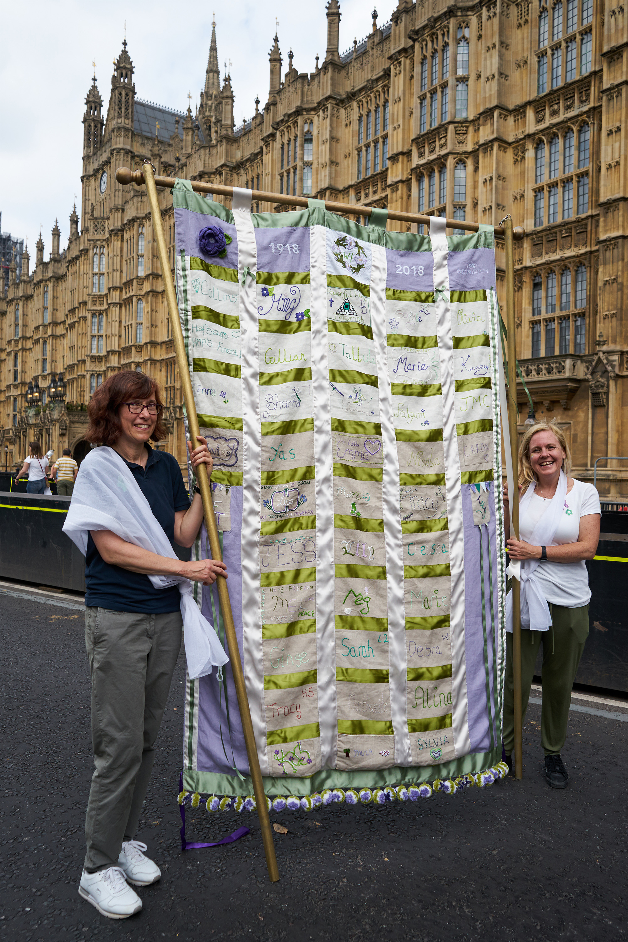 Two students pose with a seven-feet tall banner outside the Houses of Parliament.  The banner is green white and purple cloth in panels, modelled on the 20th century suffragette Holloway prison banner, embroidered with names of prisoners from Downview.