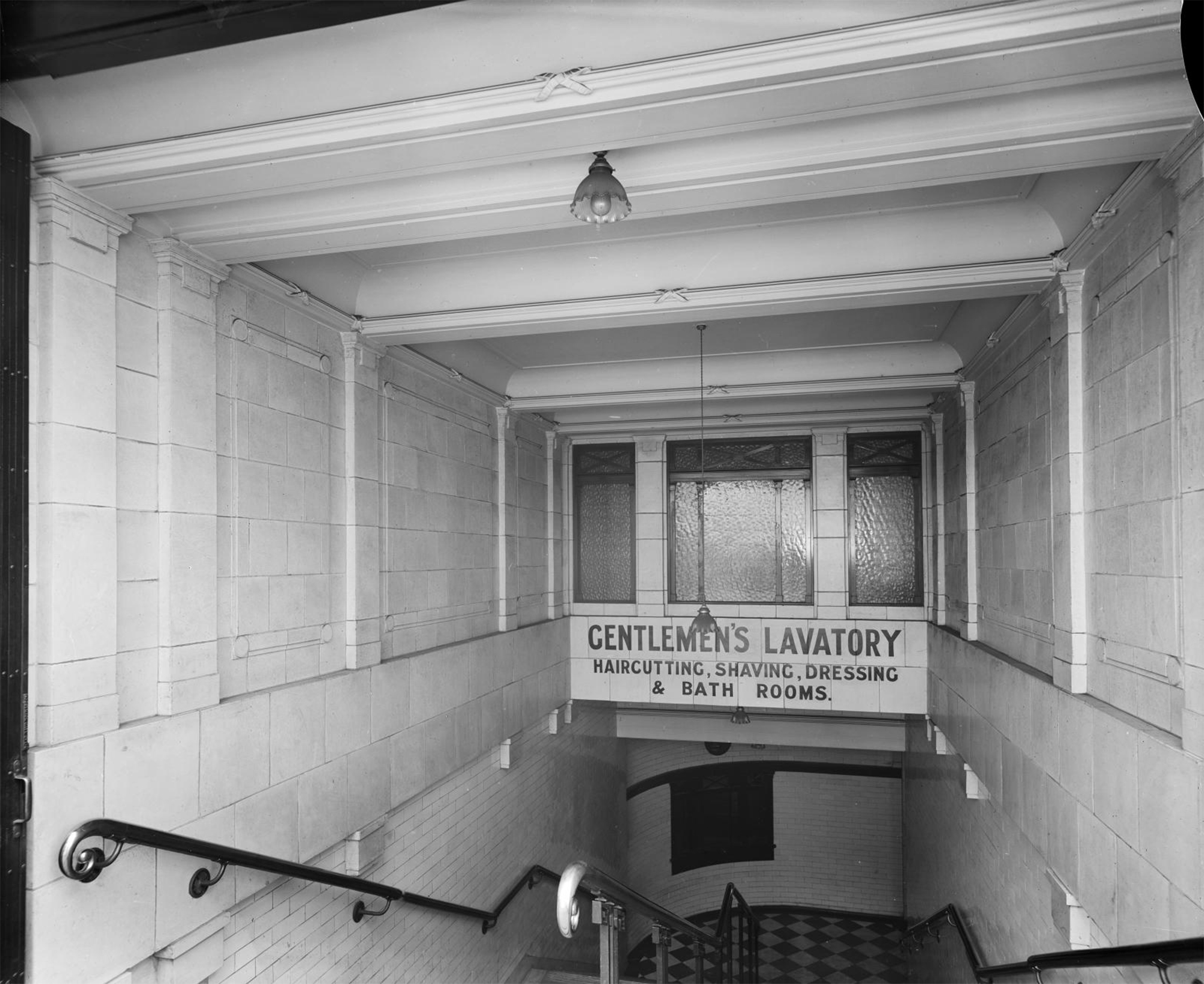 The view looking down a flight of stairs to subterranean gents' toilets. The walls are covered in white tiles.. A sign beneath a window, part way down the stairs reads: ‘GENTLEMEN’S LAVATORY, HAIRCUTTING, SHAVING, DRESSING & BATH ROOMS.’