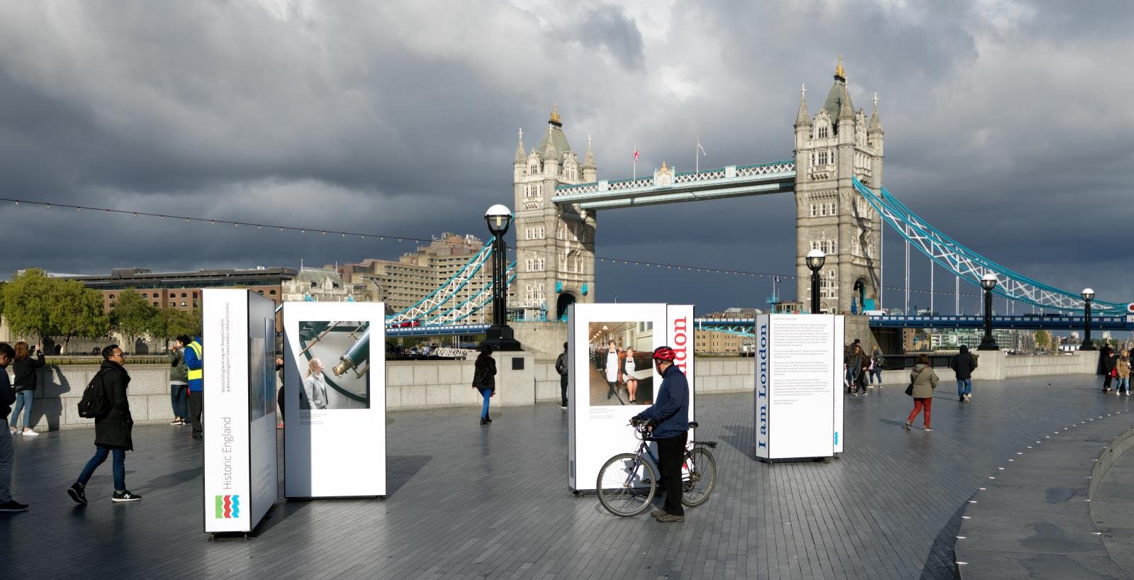 Tower Bridge, London with pedestrians and cyclists in the foreground stopping to look at Historic England's I am London exhibition panels in a pedestrianised area on the south bank.