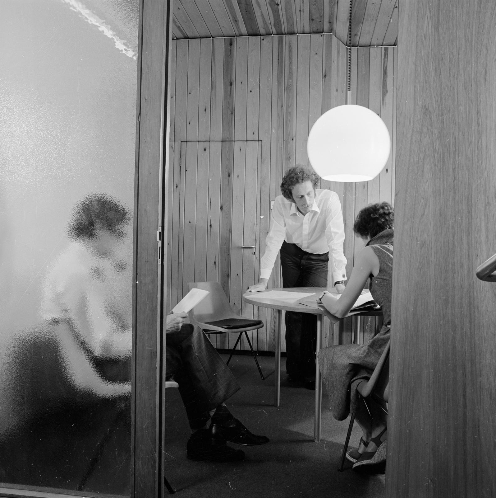 1975 office scene - black and white photo of three people around a meeting table in a timber clad room in the offices of the architects austin smith lord - as seen through a doorway.