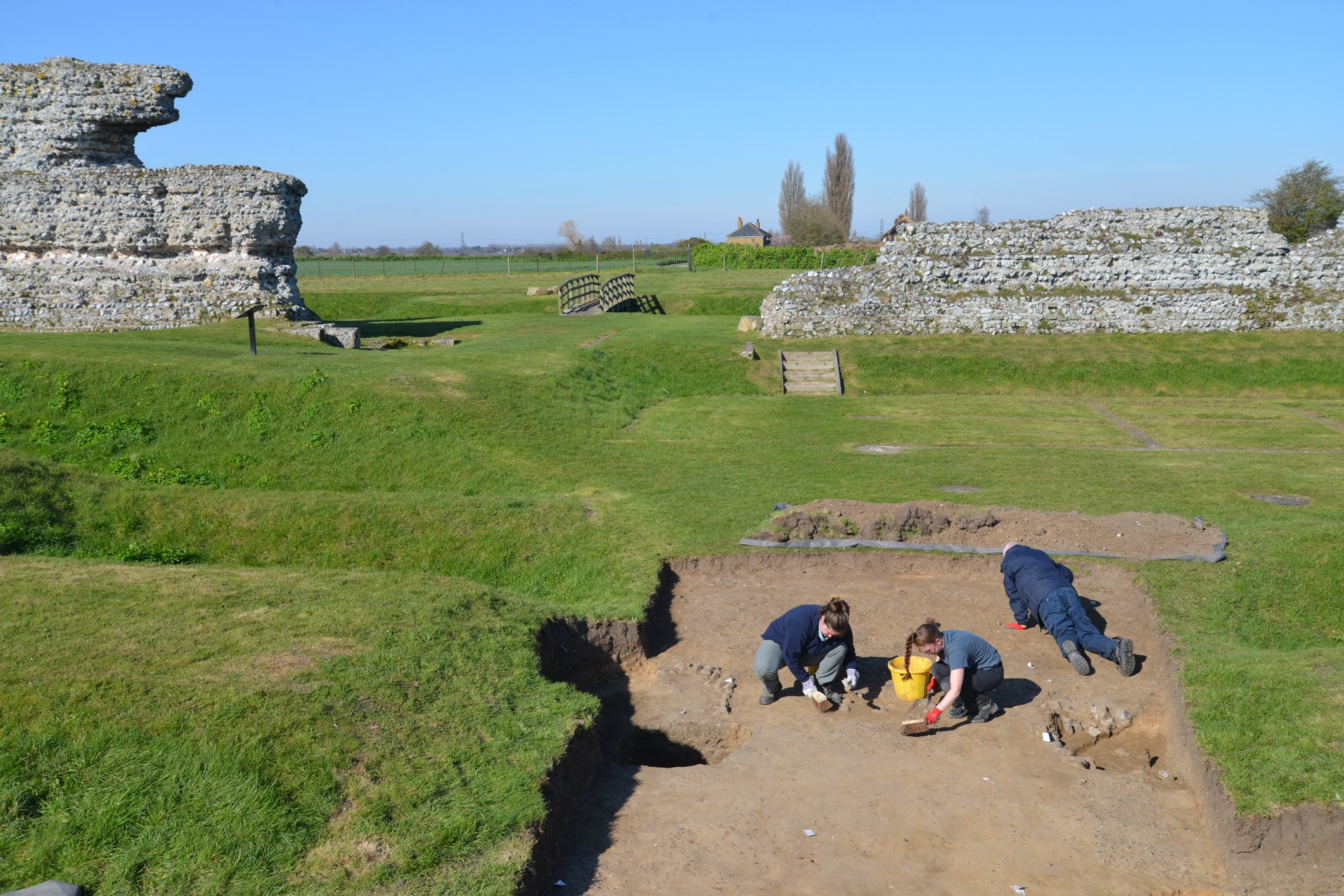 Archaeologists excavating features with upstanding ruins of Roman walls in the background.