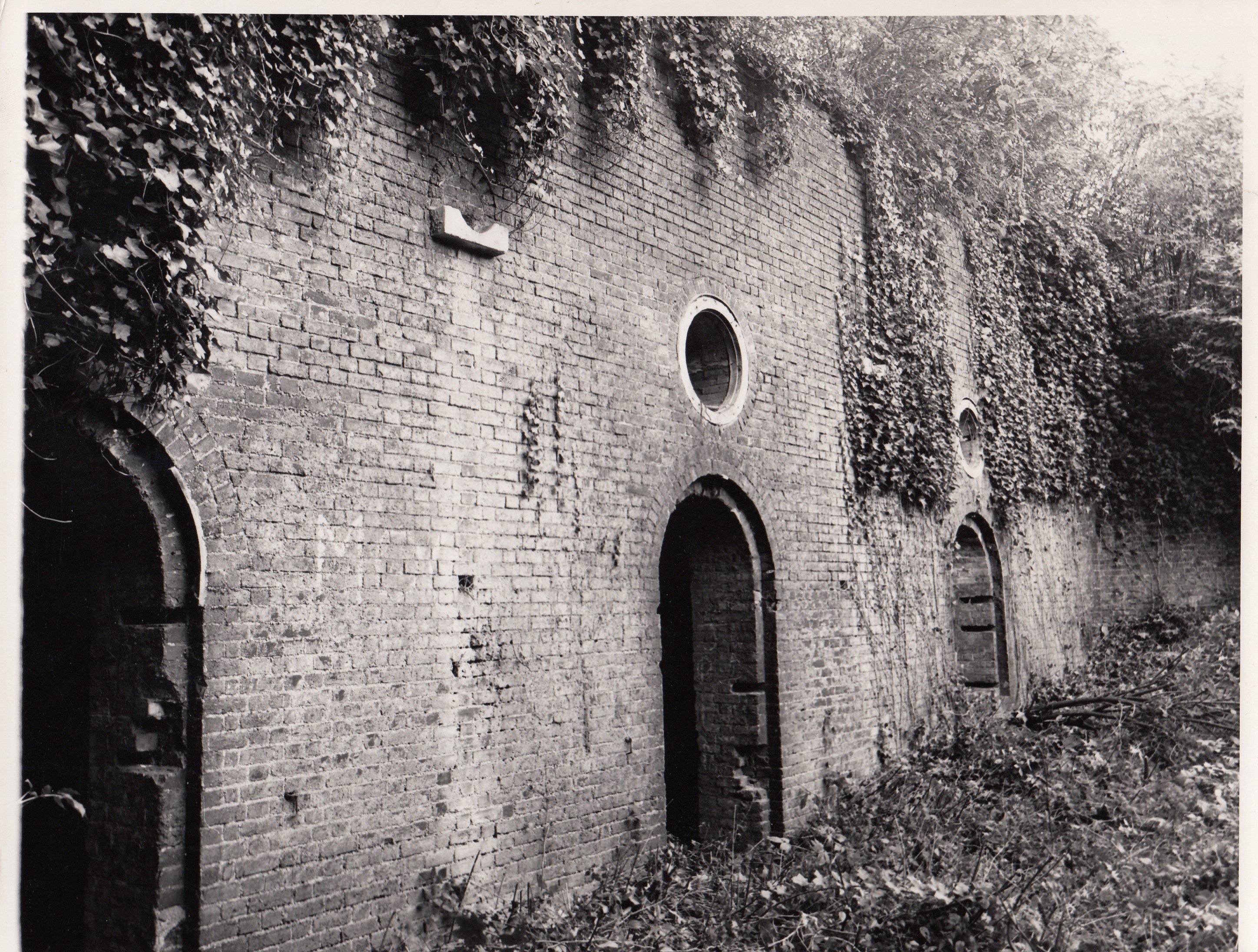 Black and white photo of high brick casemates with arched openings at ground level and circular windows.