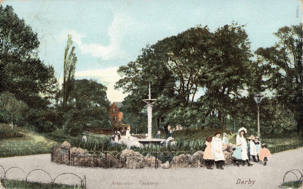 A group of women and children are gathered on a track in the right foreground, next to a fountain centre. There are trees in the background.