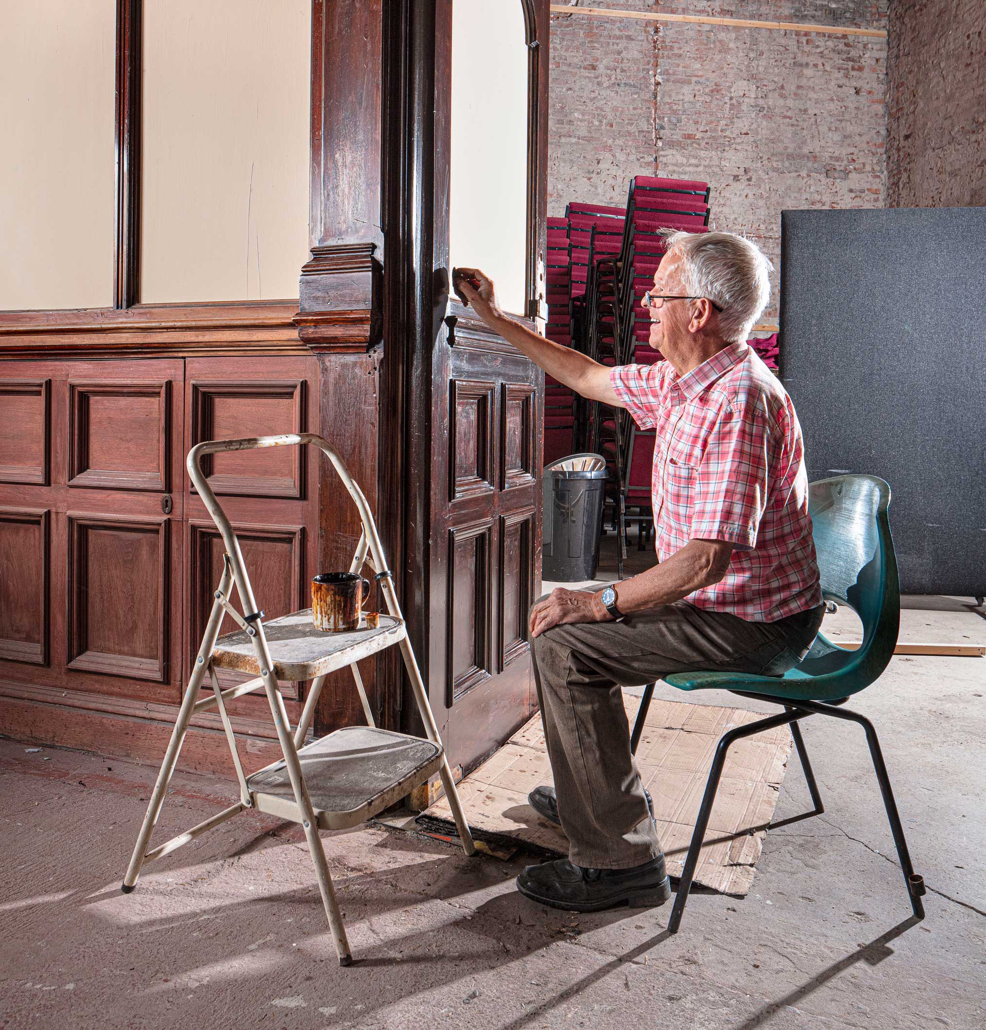 An older man seated in front of historic wood paneling which he is restoring.