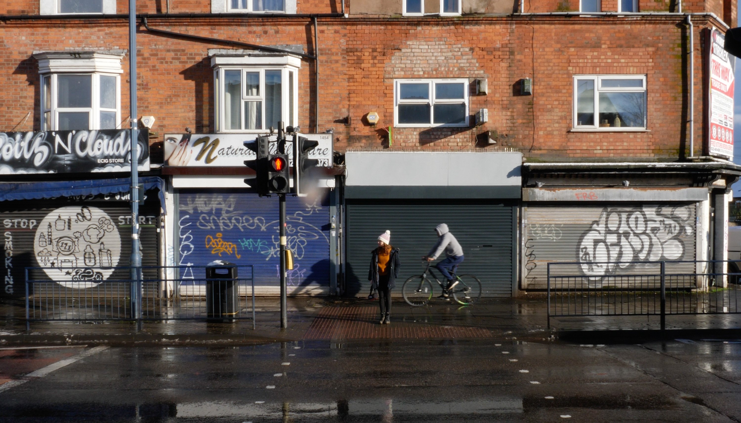 Woman stands waiting at a pelican crossing as a cyclist passes behind her on the pavement. Behind her along the street, shops are closed and most of the shop front security shutters have graffiti.