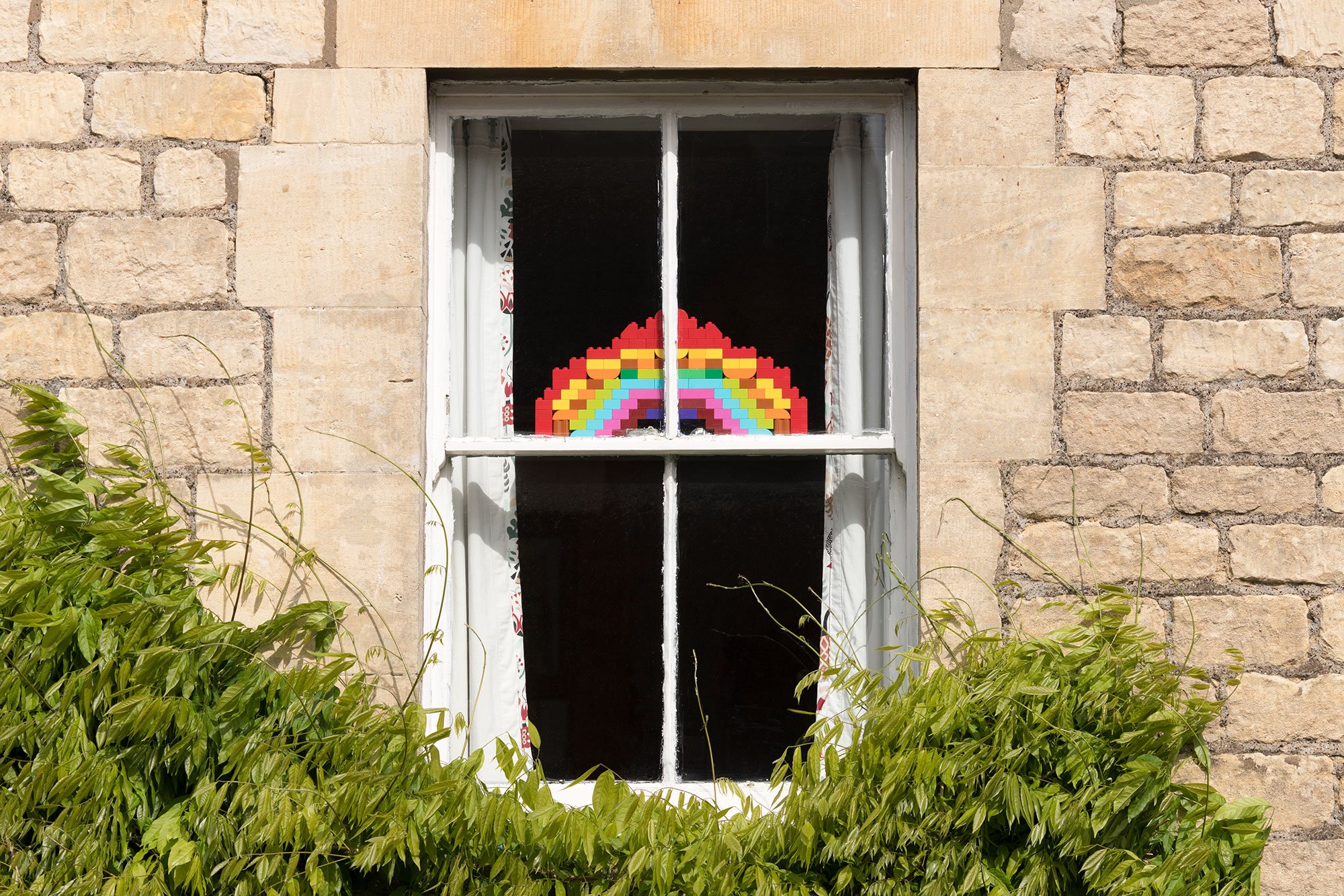 Photograph shows a view from a street of a window in a Cotswold stone house, in which is placed a rainbow made of lego.