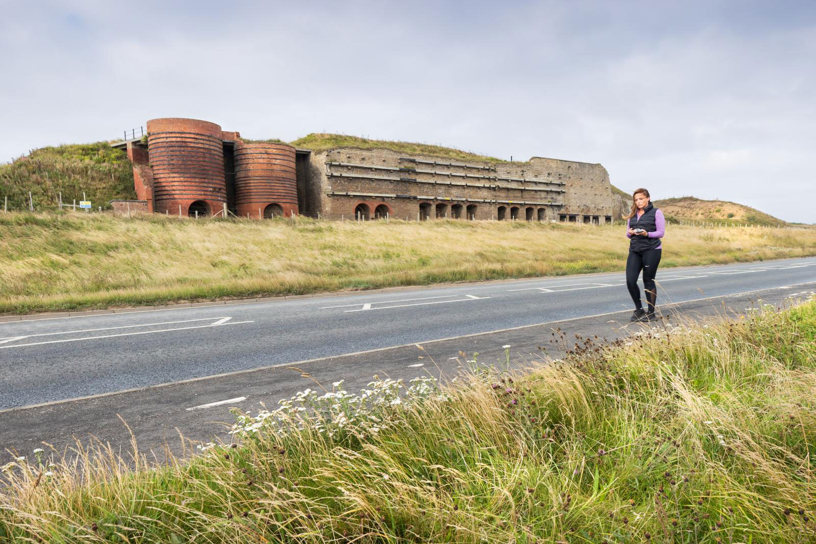 Stone- and brick-built battery structure with horizontal reinforcing timbers with a series of ground-level arched openings to the lime kilns it houses. Dune grass grows in front of it and grassy banks protrude above and either side of the structure.