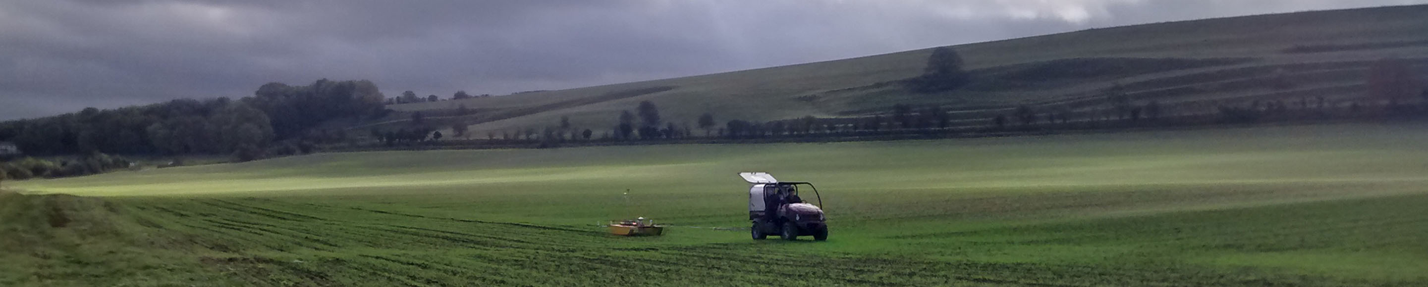 Colour photograph of buggy pulling a "sledge" across a field with germinating crop under a threatening sky heavy with clouds