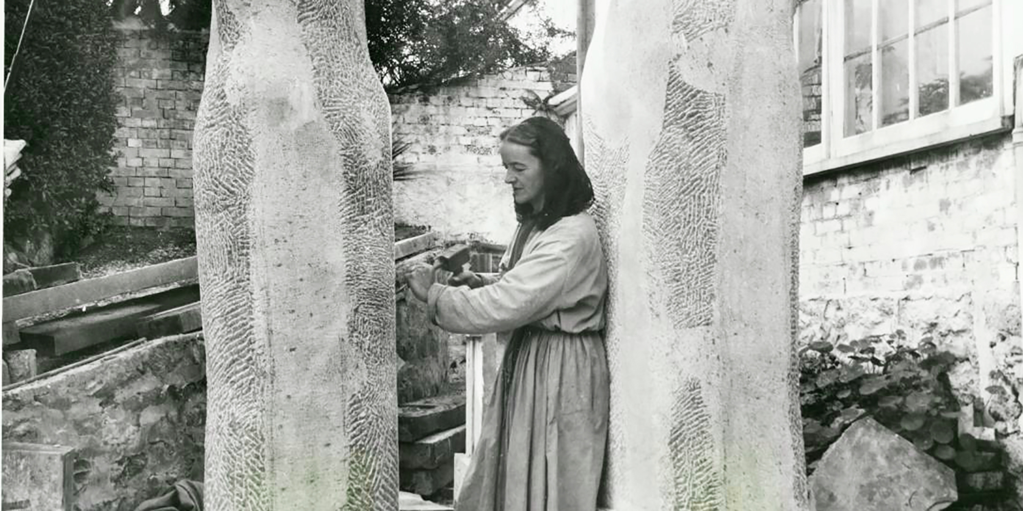A woman with a mallet and chisel carves at one of two large upright blocks of stone.