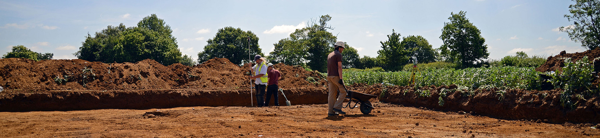 Photo of wide view of excavation trench with spoilheap at rear. In the trench is a man with a wheelbarrow and two surveying