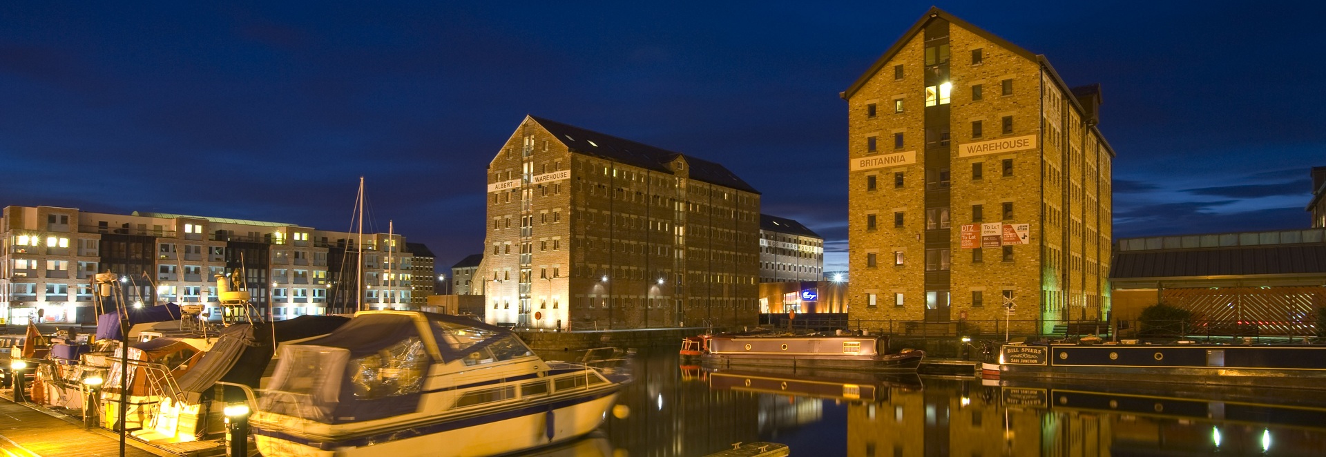 Night-time at Gloucester docks. The street lights pick out boats in the foreground. Converted warehouses stand in the background.
