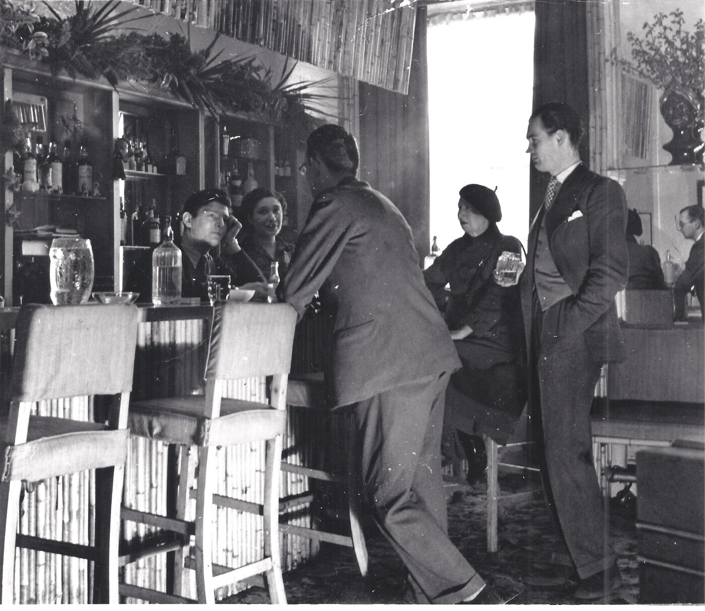 The photograph shows Ian Board, Colony Room bartender and later proprietor (seated behind bar), Carmel Stuart, Muriel Belcher's lover (seated behind bar), Nina Hamnett, artist and bohemian (seated at bar)