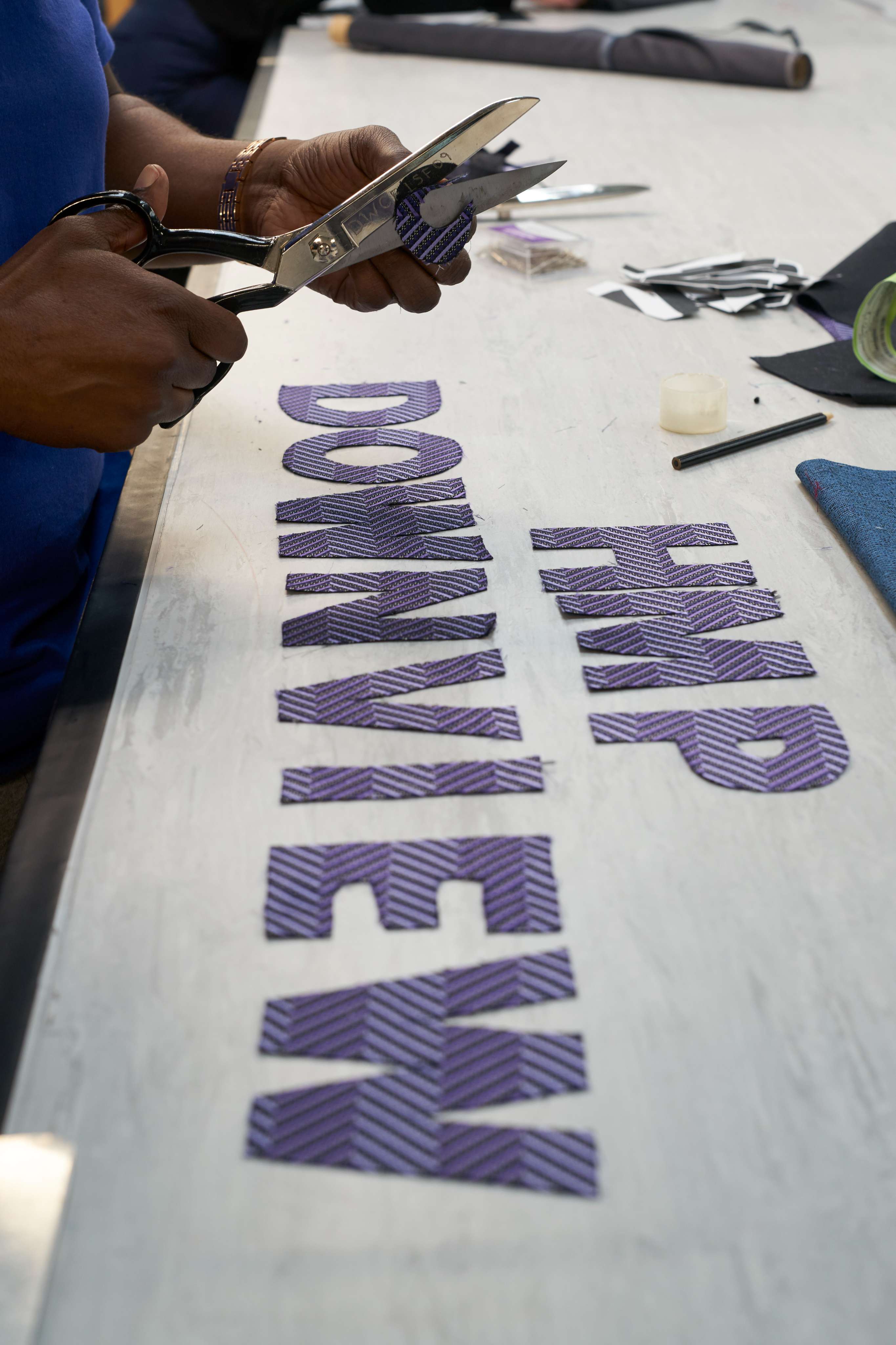 A pair of hands cuts the letters 'HMP DOWNVIEW' out of purple fabric.