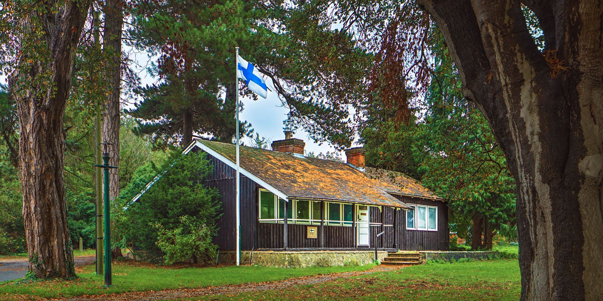 A low wooden building in parkland, with orange leaves on the roof and a Finnish flag flying in the foreground.
