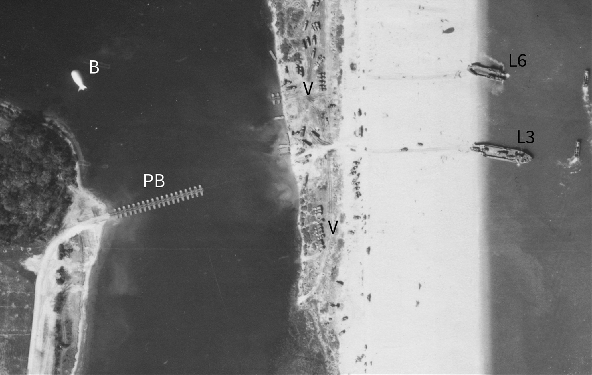 Detail from a black and white vertical aerial photograph, showing a pontoon bridge being constructed on a stretch of water between a landward road and a narrow strip of beach. Two landing craft are stationary at the edge of the beach. Vehicles disembark from one of them. Smaller landing craft manoeuvre on the sea close to them. Various vehicles are on the beach and on land between the beach and the pontoon bridge. The letters and numbes 'PB', 'B', 'V', 'L3' and 'L6' applied to the image mark locations.