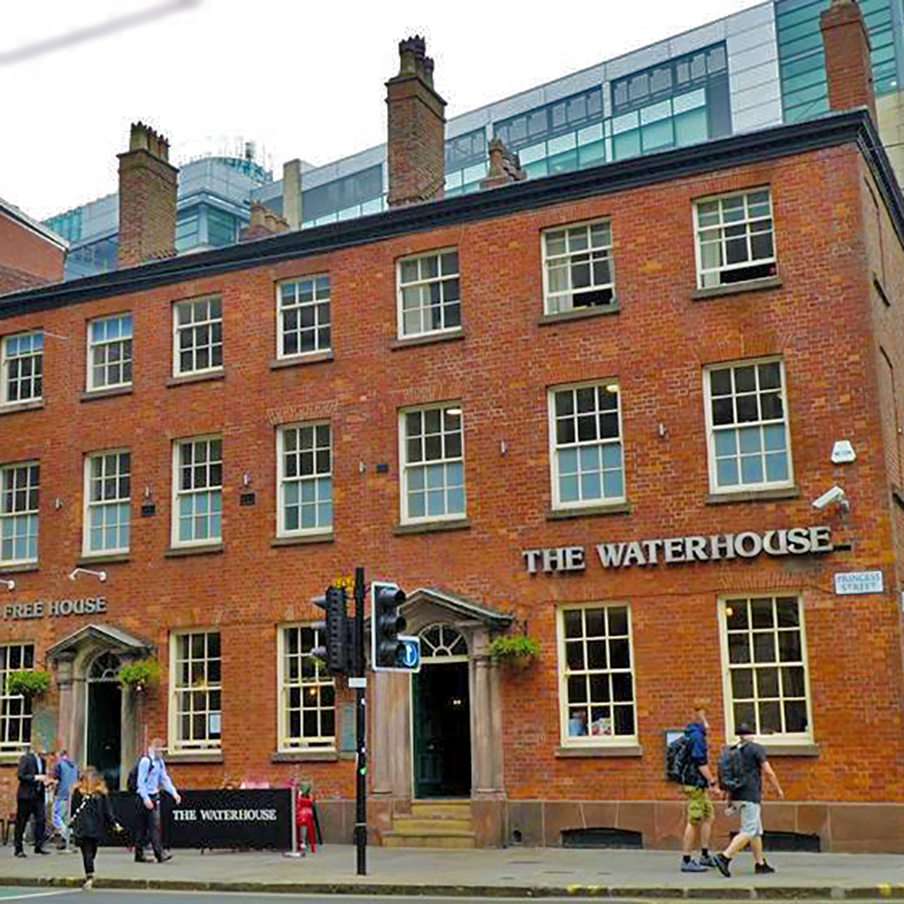 Pedestrians pass in front of a broad-fronted 3-storey red-brick town building with signs saying "The Waterhouse", "Free House" and "Princess Street".