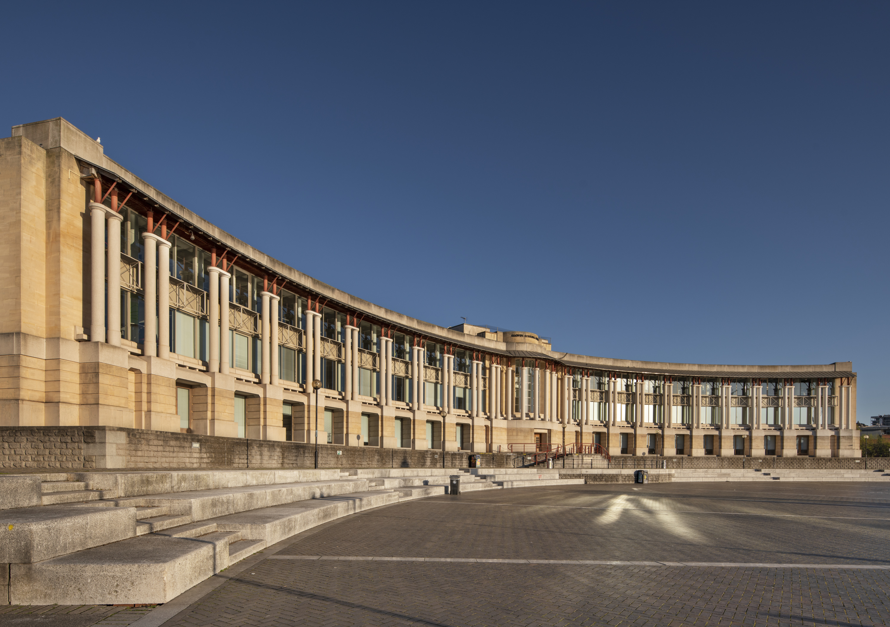 A grand, modern building with a curved frontage and colonnade overlooks a paved public space