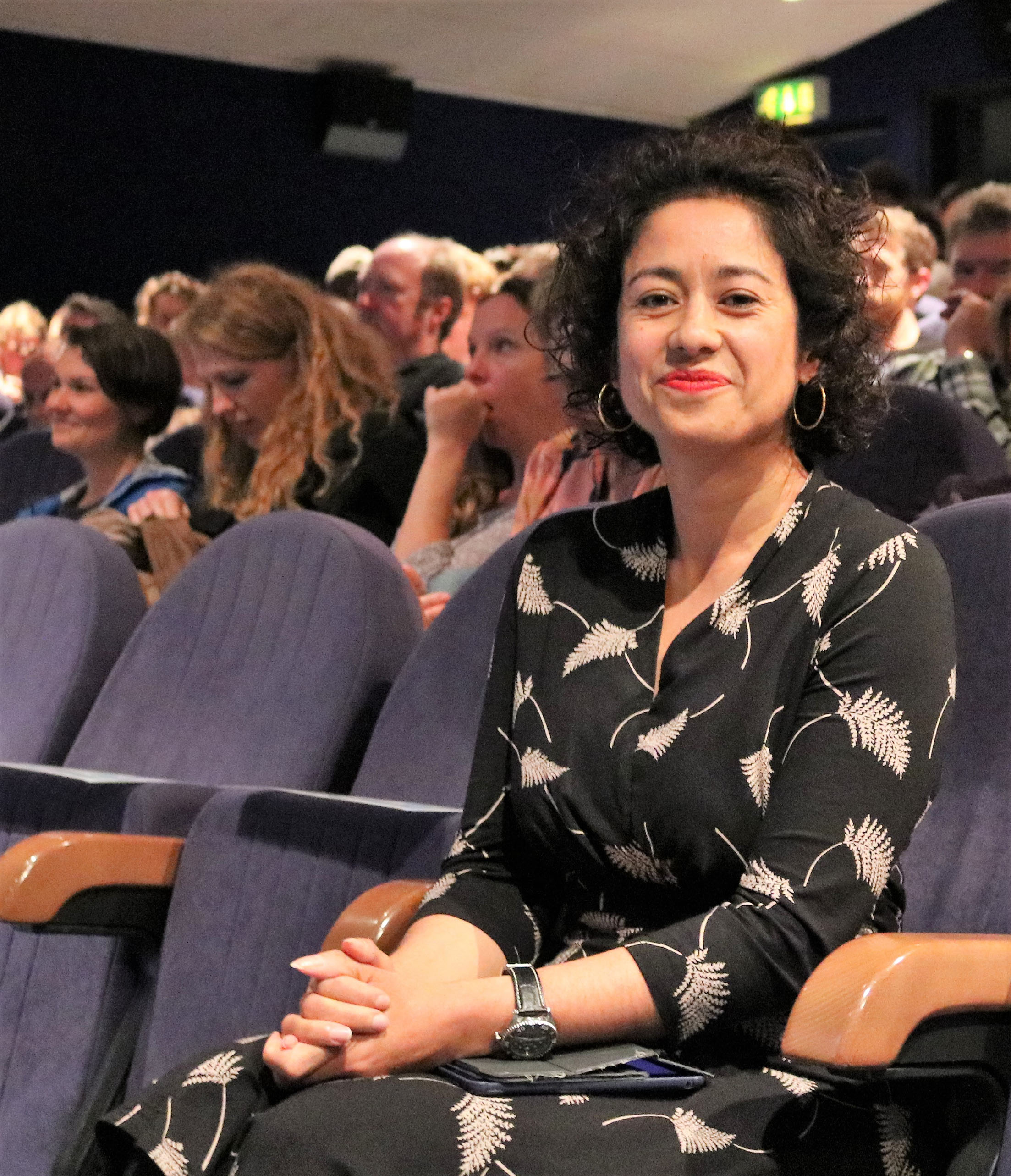 A portrait photograph of Samira Ahmed sitting in a blue auditorium seat, with other people sitting in the seats behind her