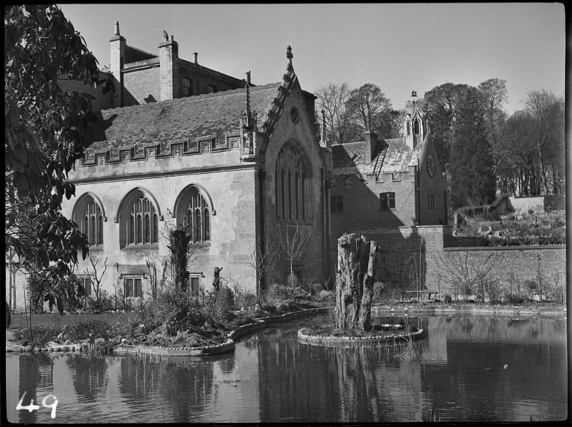 Black and white photograph of part of a country house, viewed from the other side of an ornamental pond. In the pond is a circular island with an elongated tree stump. On the far side are planted borders behind which rises a Gothic-style chapel building. Rising behind are other elements of the house.