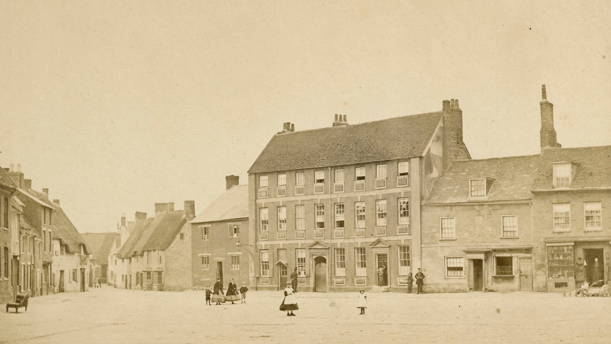 Archive sepia photograph looking across a wide paved space with houses and shops both on the far side and lining a road leading away to the left. People in period costume, mostly children, are standing looking to camera.