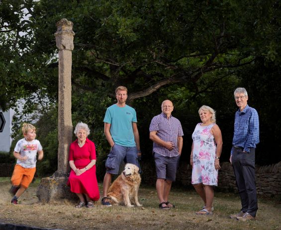 A colourful group of six people and one dog pose next to the Village Cross; an approximately-three meter high stone monument. 