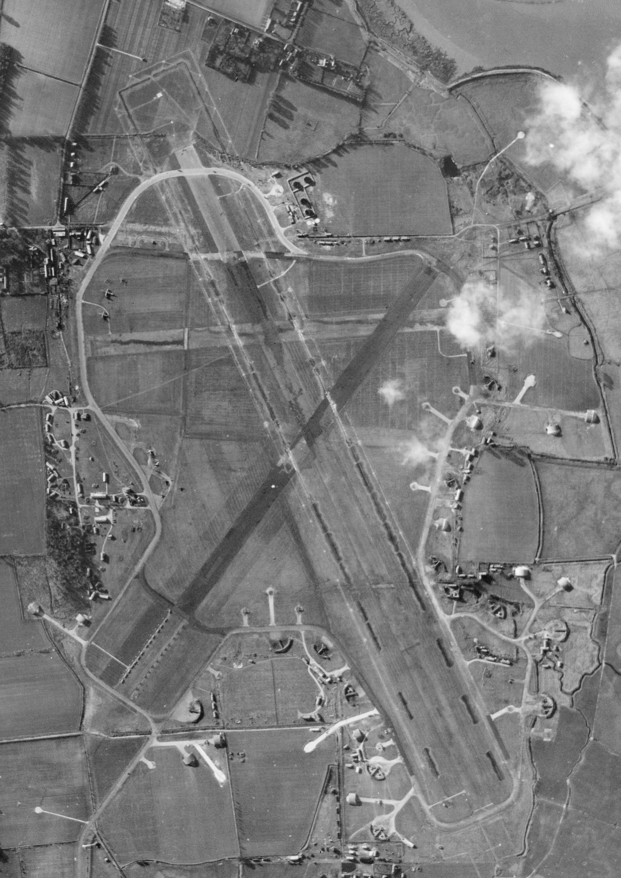 A black and white vertical aerial photograph showing an airfield with three runways. Dispersal areas with hardstands protrude from an irregular perimeter road.