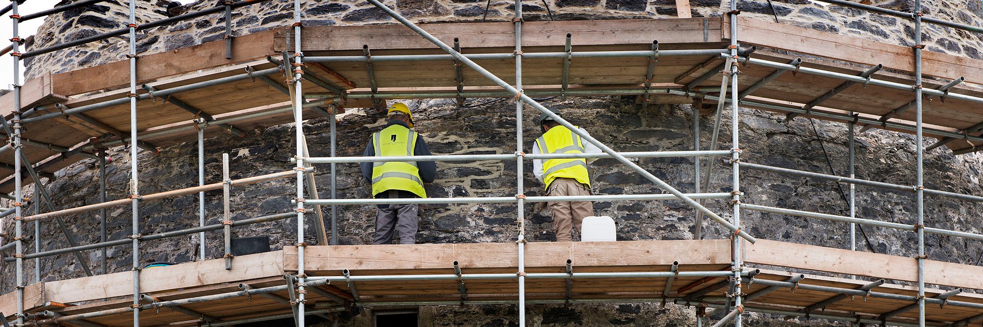 A photograph from behind of two individuals within scaffolding working on a stone wall.