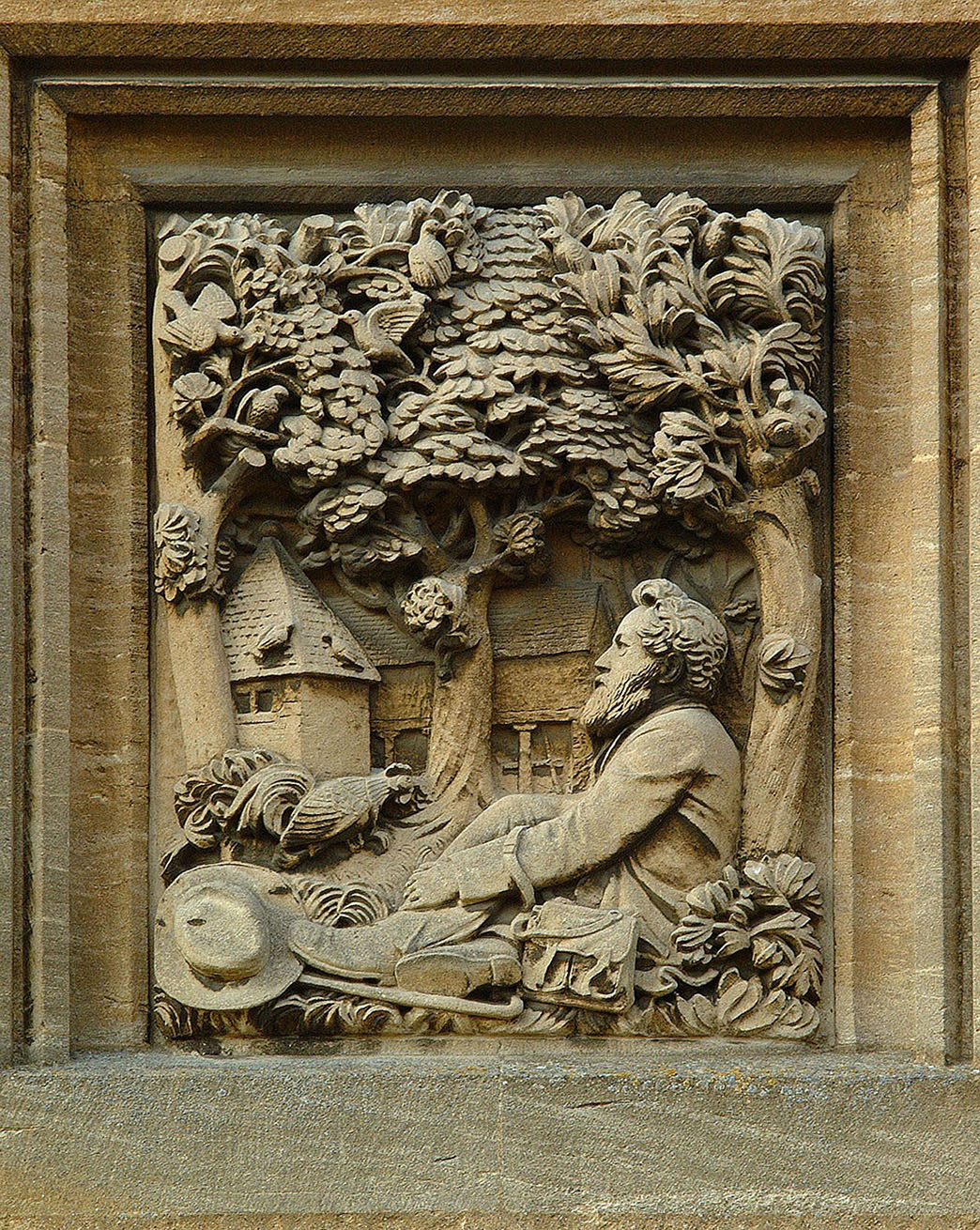 Stone carving on one of the cottages in the village showing William Morris sitting under a tree at Kelmscott with the Dovecote and farm barns behind him.