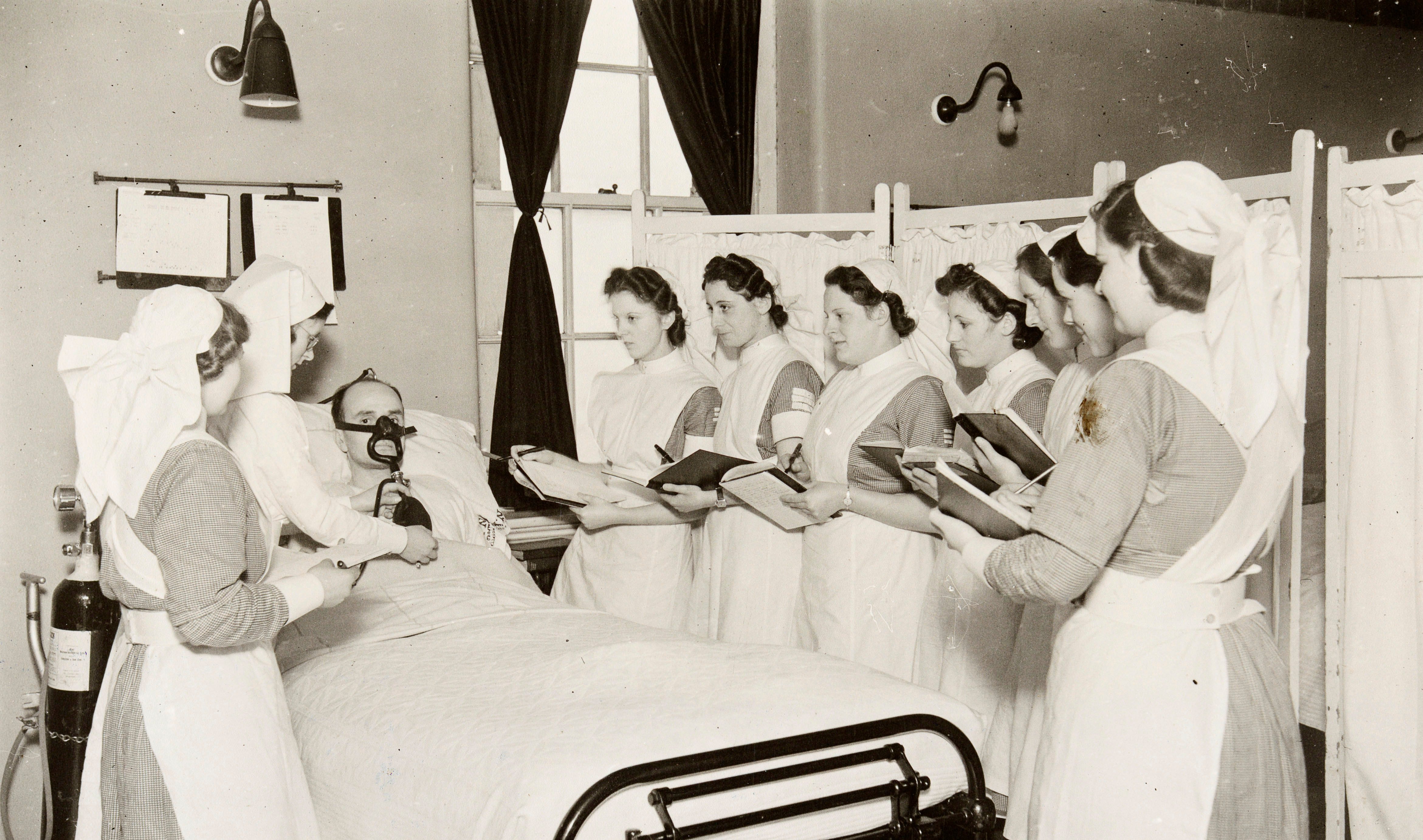 05 Dec 1940. The caption on the reverse of the photograph reads: “Hope Hospital, Salford. Picture shows the Sister Tutor, Sister F. E. Knowles, giving a class of nurses who are taking the Preliminary State Examination in February a demonstration in the ward on the B.L.B. oxygen mask.” Source: Historic England Archive MED01/01/1358