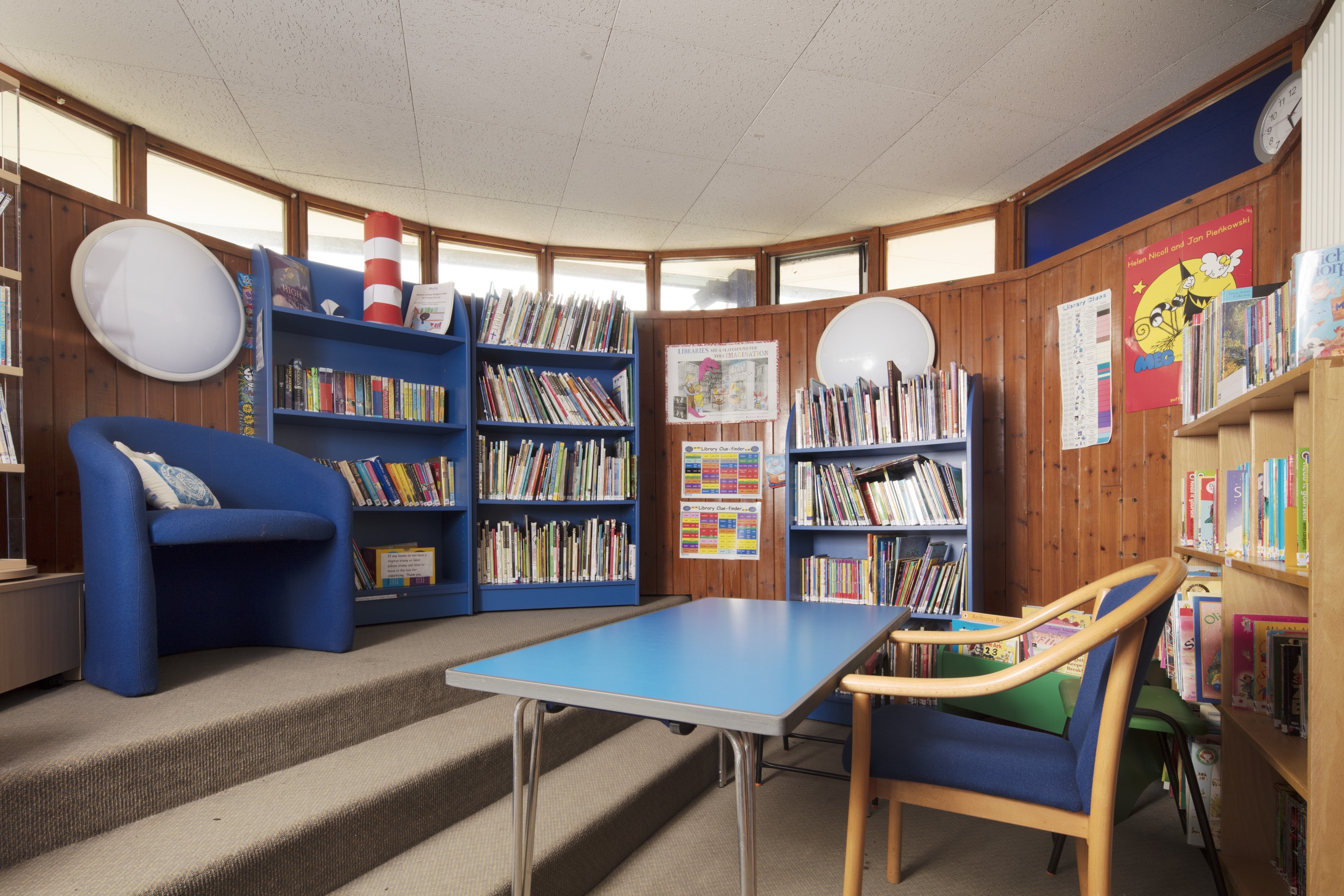 Photo of the interior of a school classroom with bright blue bookshelves and seats. The walls are wood panelled with small windows at the top.