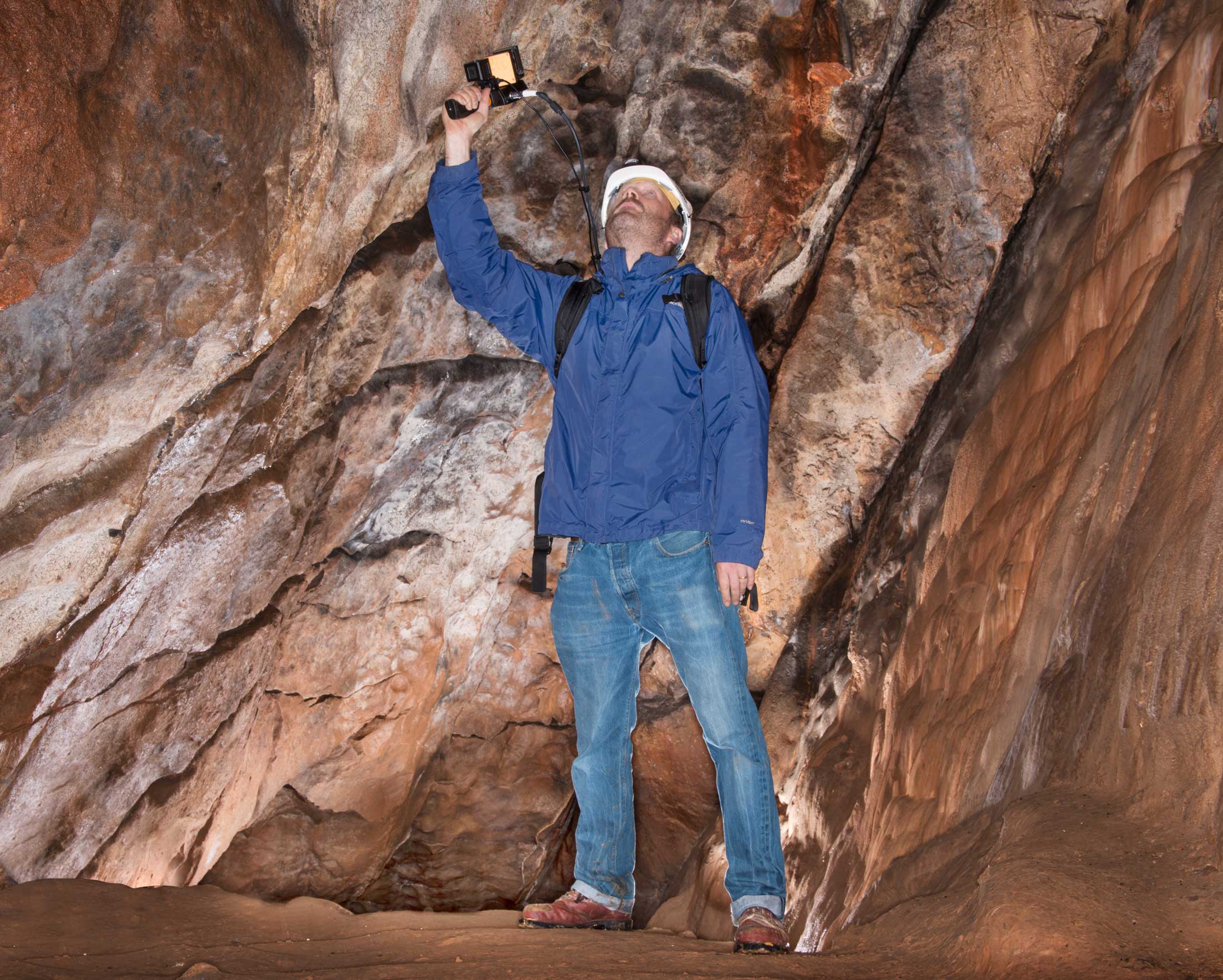 Man standing in a cave wearing outdoor clothing and hard hat, holds a scanning device above his head and looks up towards the roof of the cave.