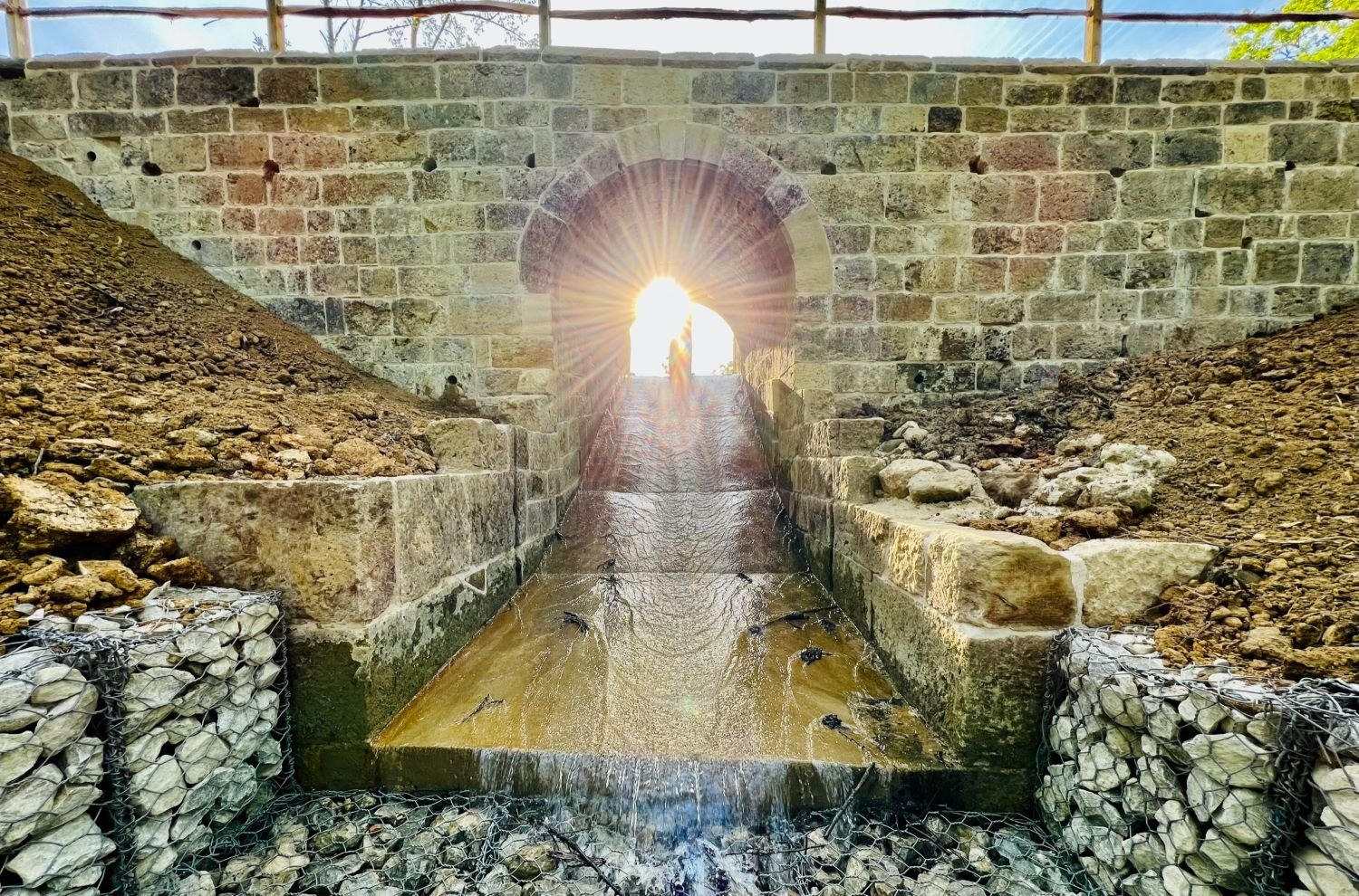 Stone wall with a central archway over a water sluice. The sun is shining through the arch.