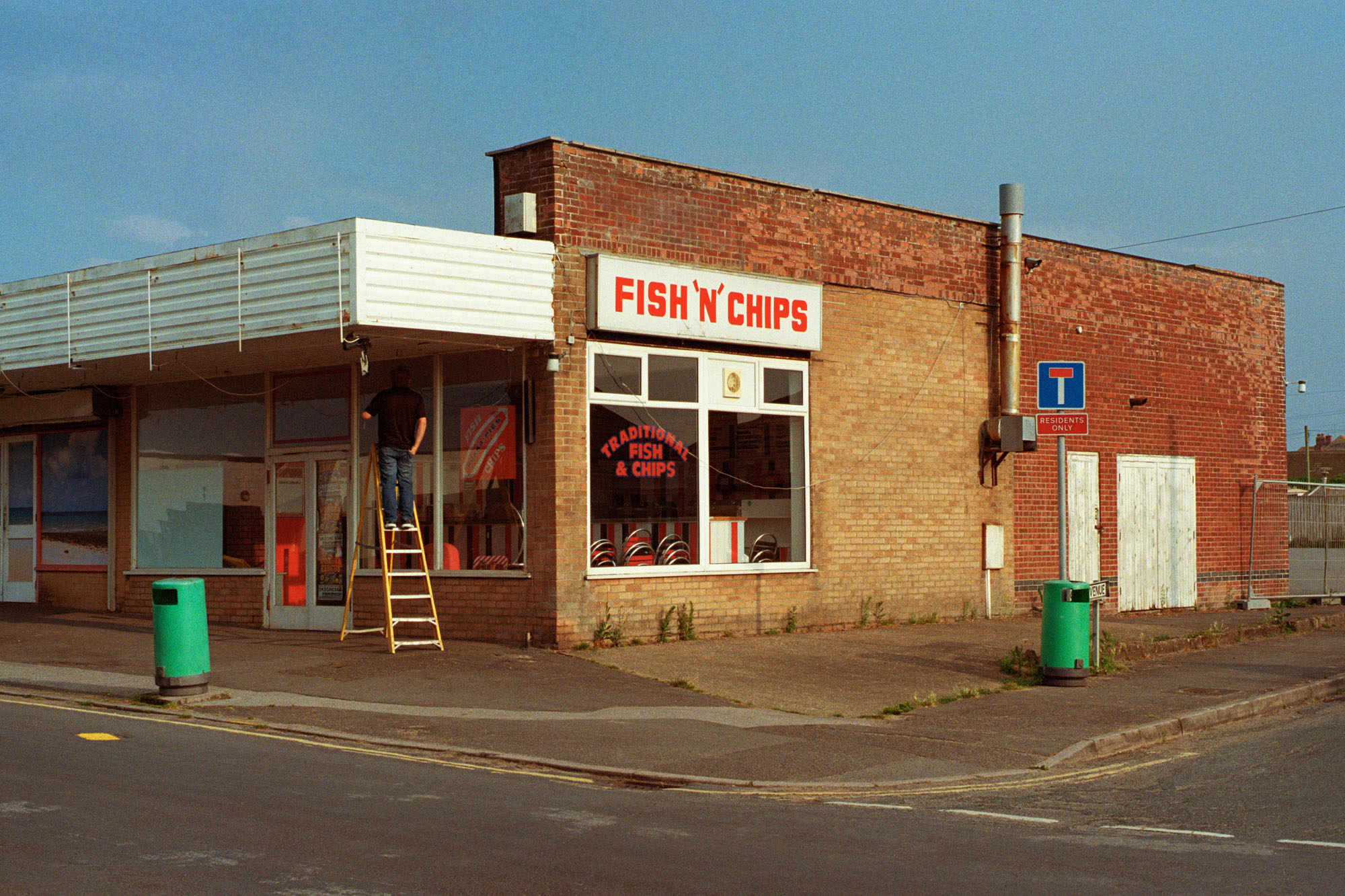 A colour photo a single-storey commercial building on the corner of two streets. The building is faced in brick, The front includes a deep overhang. A sign above a large window reads: 'Fish 'N' Chips'. A person on a step ladder works beneath the overhang. On the pavements to the front and side of the shop are green rubbish bins.
