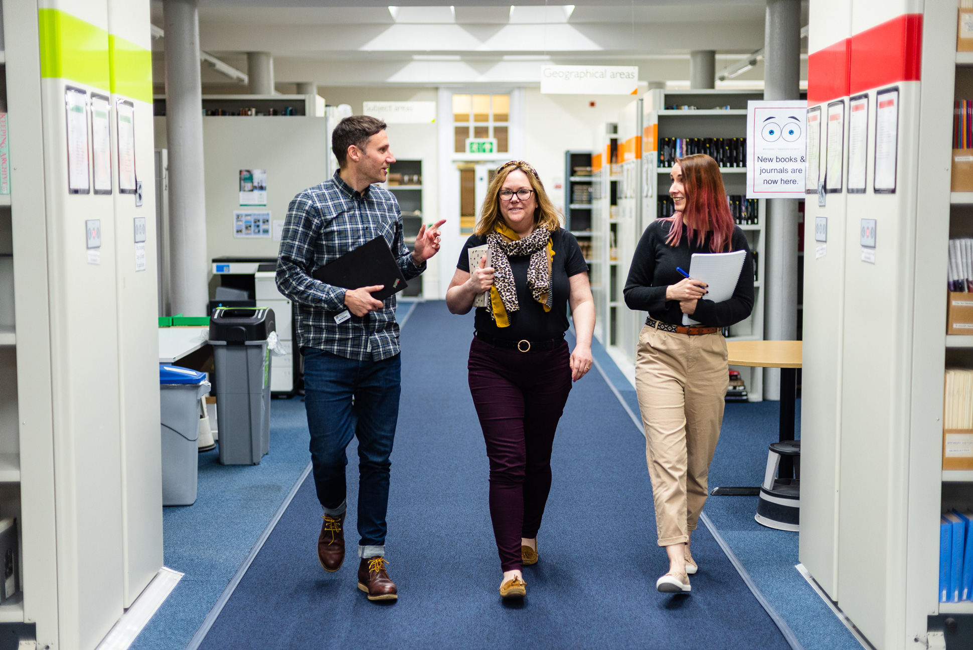 Three people walking along an aisle lined by library shelving.