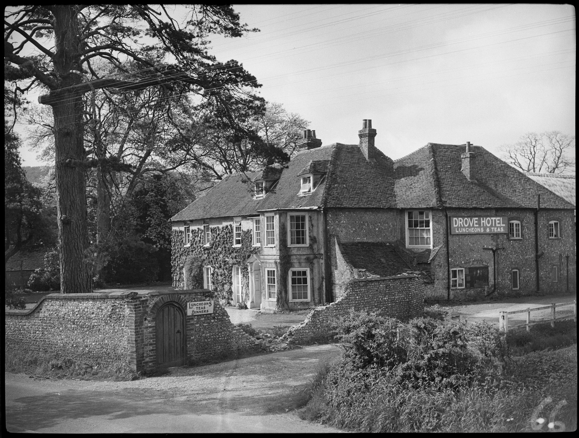 Black and white photograph of a two-storey, L-shaped house. Taken from a slightly elevated position, the view shows two sides of the building. In the foreground, damage to a wall exposes the two-storey canted bay of part of the building. A sign on one elevation reads: 'Drove Hotel' and a sign next to a door in the wall reads: 'Luncheons Teas Dinners'.