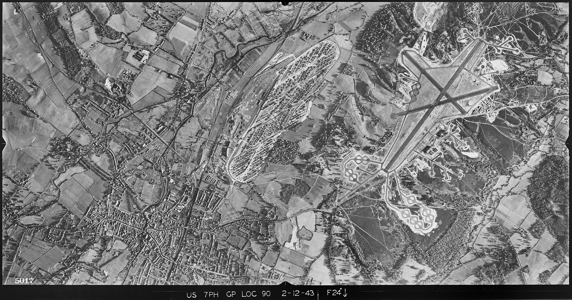 A black and white vertical aerial photograph featuring an urban area close to an airfield with three runways. Between are patches of woodland and and fields.