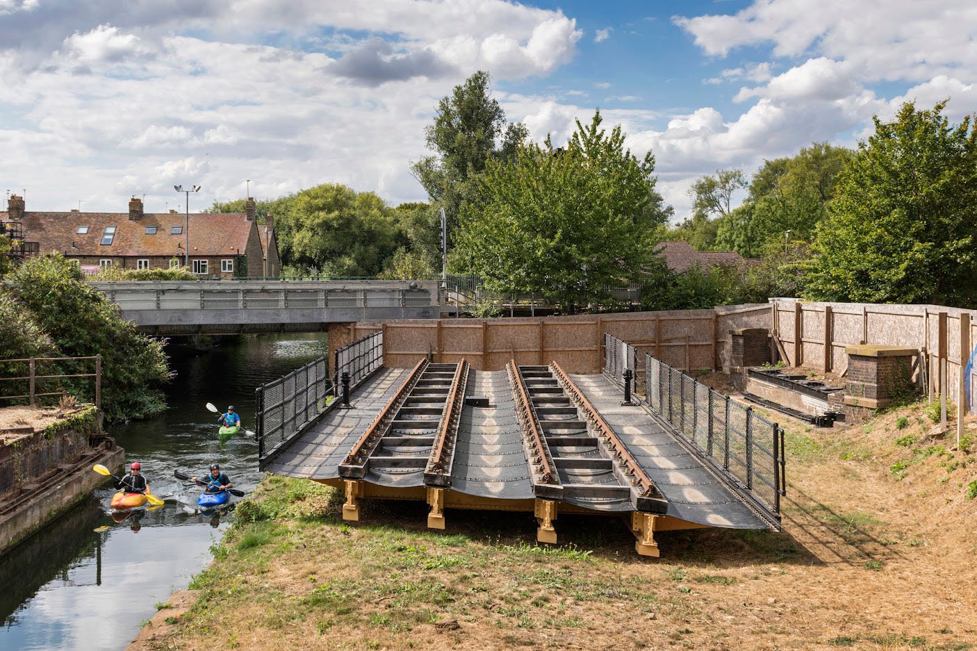 A metal structure with railway tracks sits on the bank next to a river with canoeists.