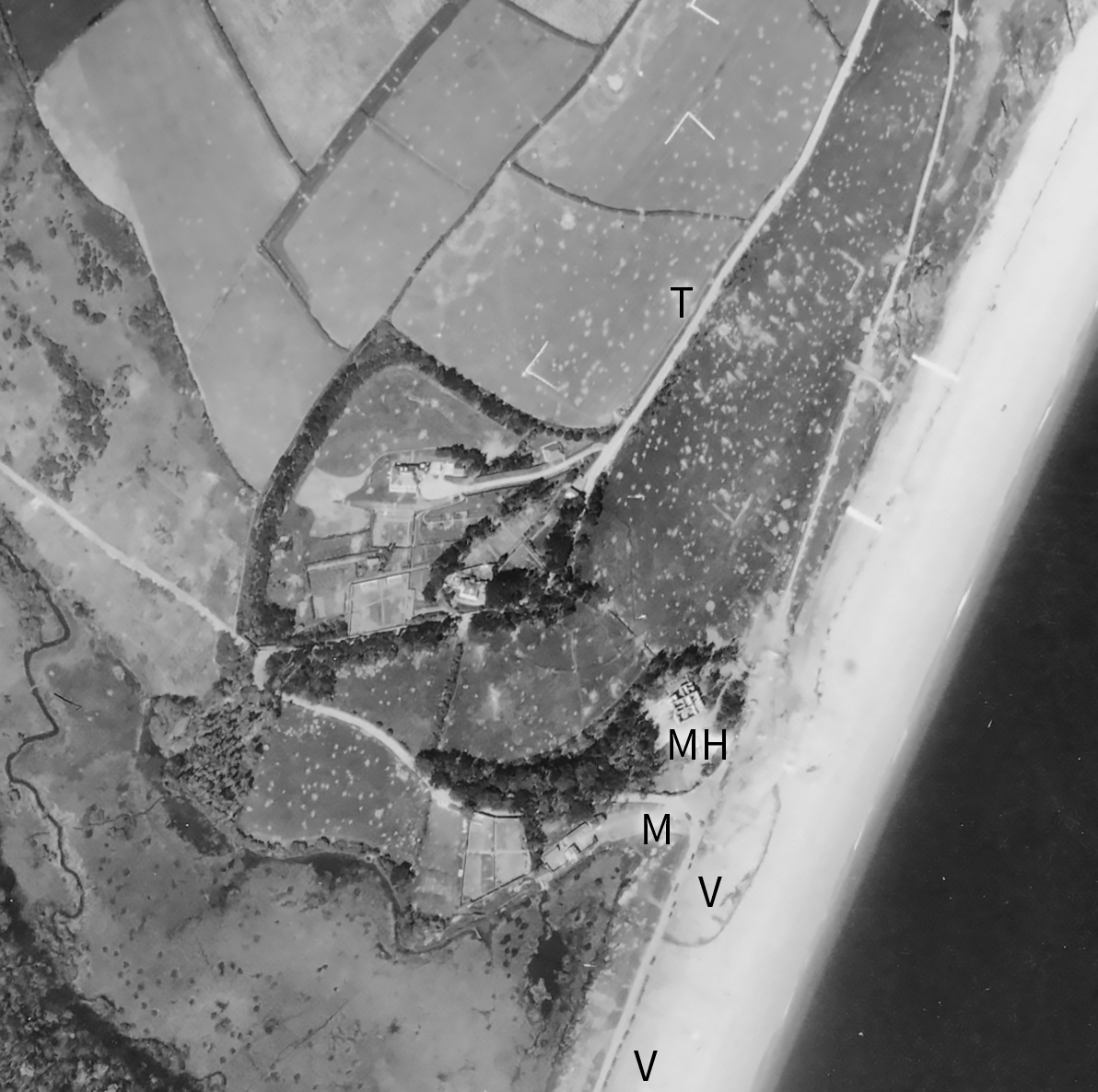 Detail from a black and white vertical aerial photograph looking down on a group of buildings adjacent to a narrow beach. The sloping hill leadingfrom the beach and the fields behind are pockmarked with shell holes. The building closest to the beach is roofless. On the sloping hill and in the fields behind are L-shaped markings, forming the corners of an incomplete rectangle. The letters 'MH', 'V', 'M' and 'T' applied to the image mark locations.